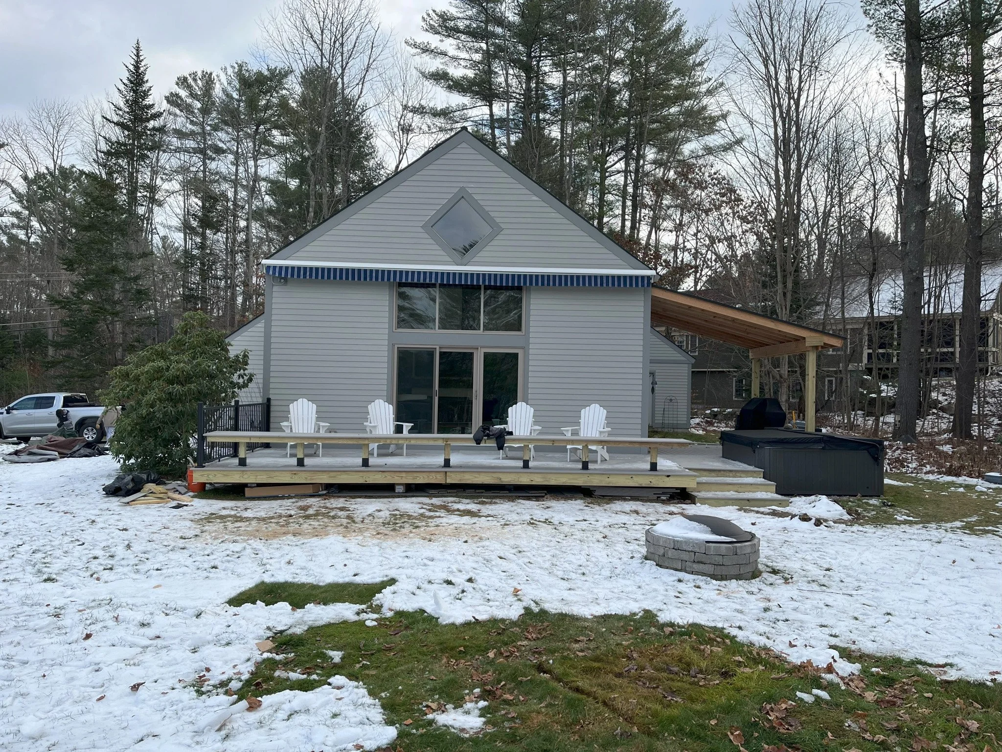 Back view of a house with a wooden deck, four white Adirondack chairs, a fire pit, and snow on the ground, surrounded by trees.