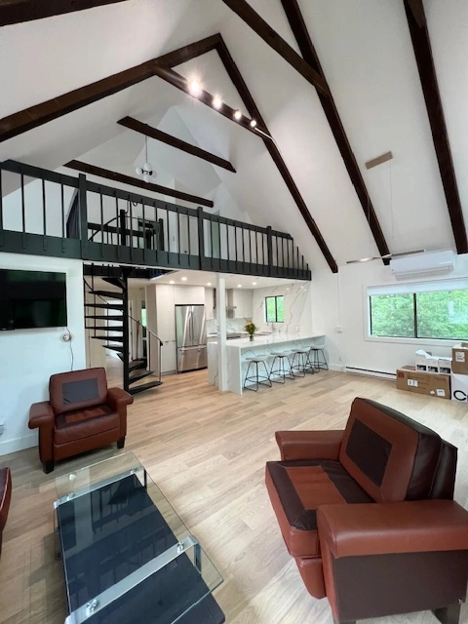 Open living space with vaulted ceiling featuring dark wood beams, a loft area with a black railing, and large windows with a view of greenery outside. There are leather armchairs, a glass coffee table, a bar area with stools, and cardboard boxes in t