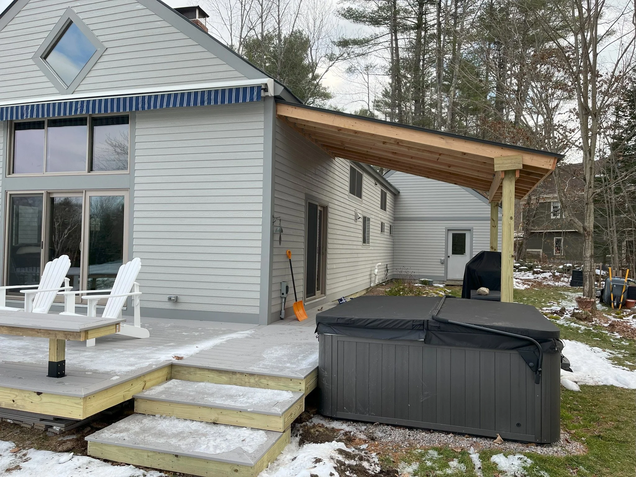 Backyard with a newly built wooden deck, two white Adirondack chairs, a hot tub, and a house with gray siding. Snow is on the ground, and the deck has small steps leading down to the grass.