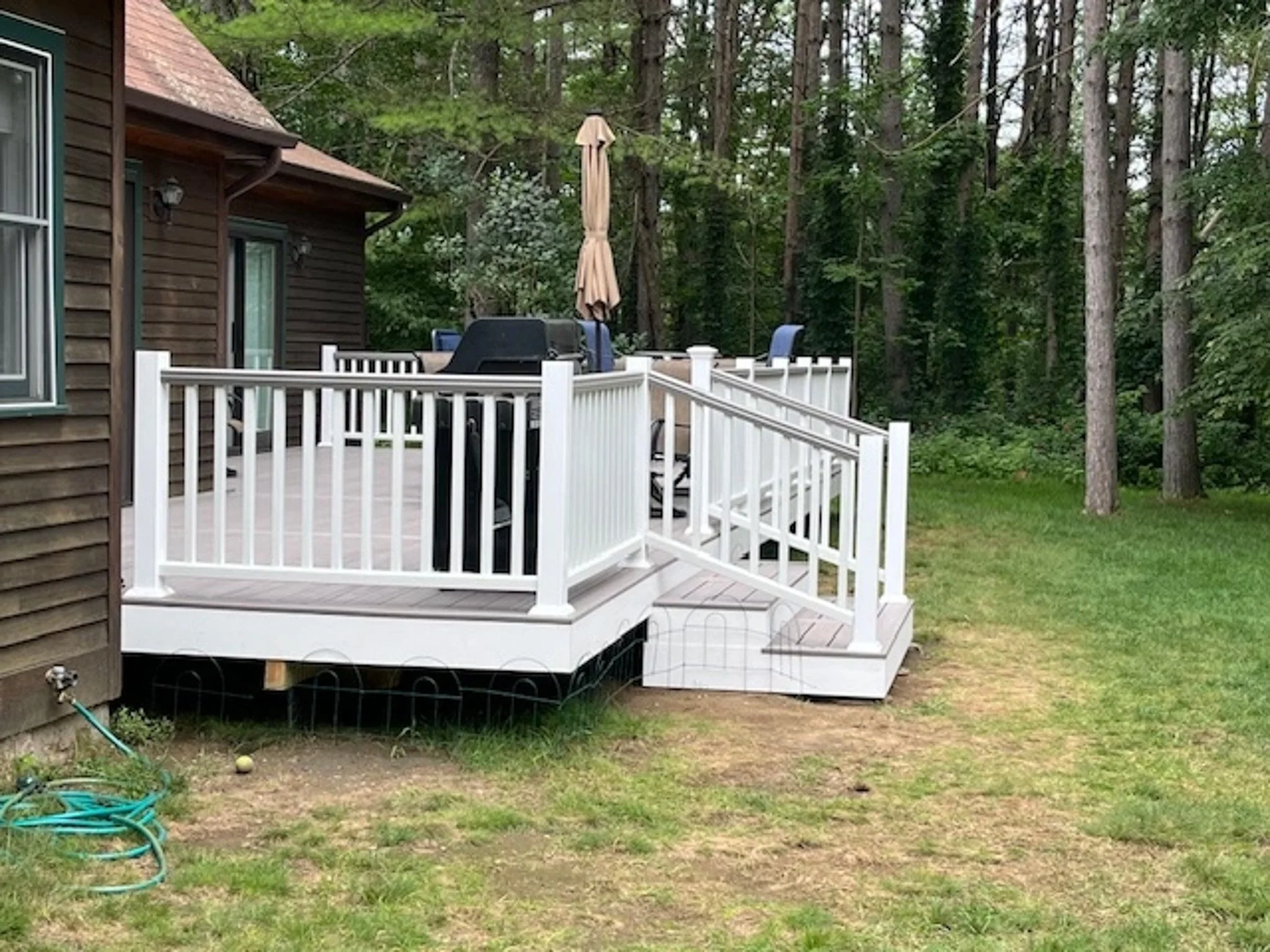 A backyard deck with white railing and stairs, outdoor furniture, a grill, a closed umbrella, and surrounded by green trees and grass.
