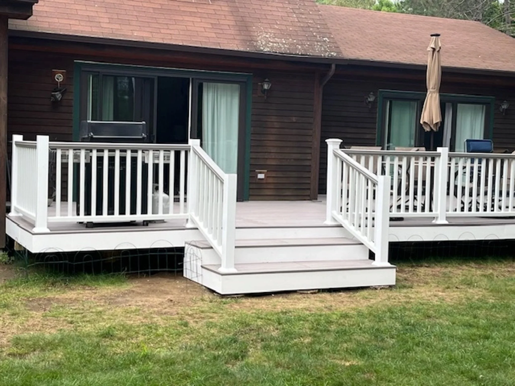 Wooden house with a large porch, white railing, sliding glass doors, and an outdoor dining area with an umbrella, set in a grassy yard.