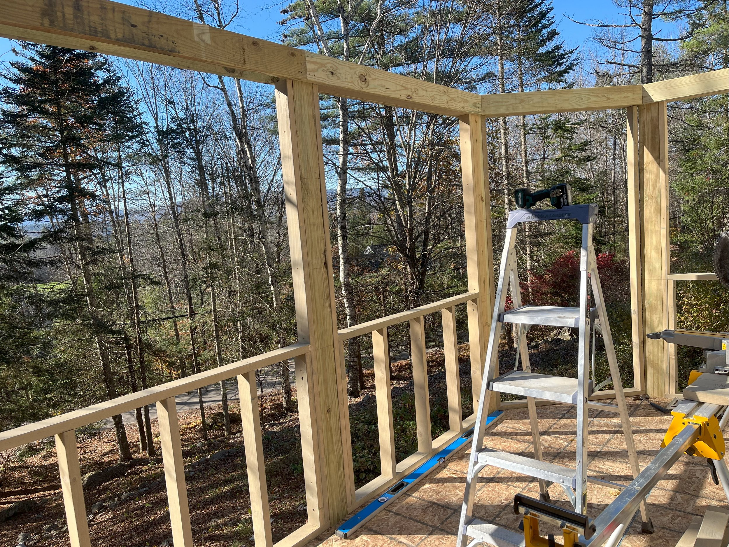 Construction of a wooden balcony or deck in a wooded backyard with ladders and tools, overlooking trees and blue sky.