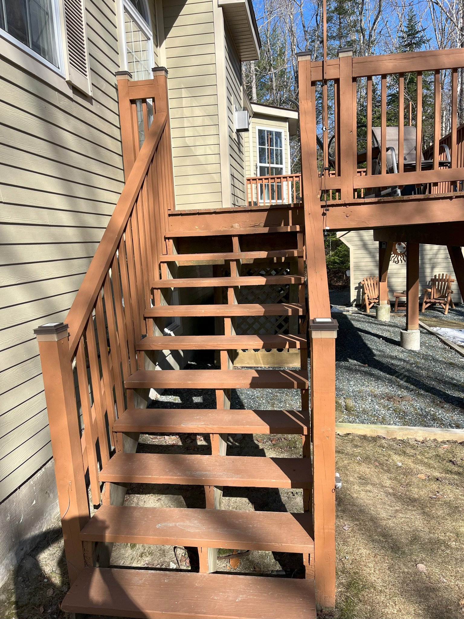 Wooden outdoor staircase leading up to a deck with outdoor furniture, adjacent to a beige house with large windows, in a backyard with trees and a gravel walkway.