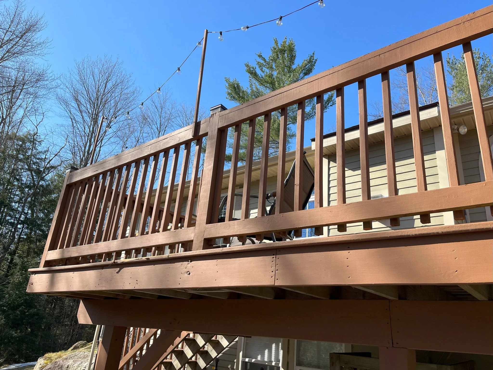 View of a wooden deck with railing, string lights overhead, partially blue sky, and trees in the background.