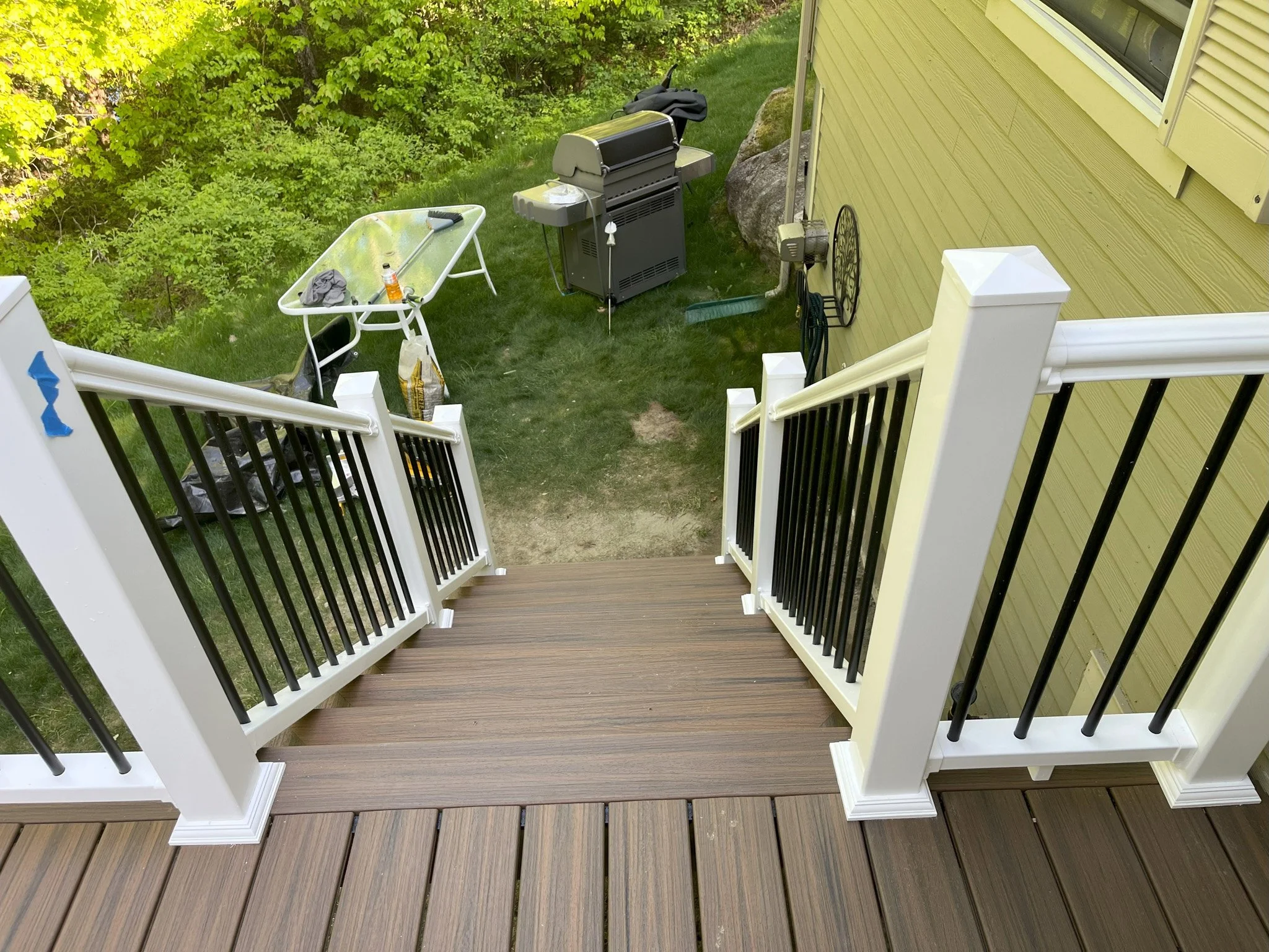 View from a deck looking down on a green yard with a yellow house to the right. The yard has a grill, a wheelbarrow, and gardening tools scattered around.