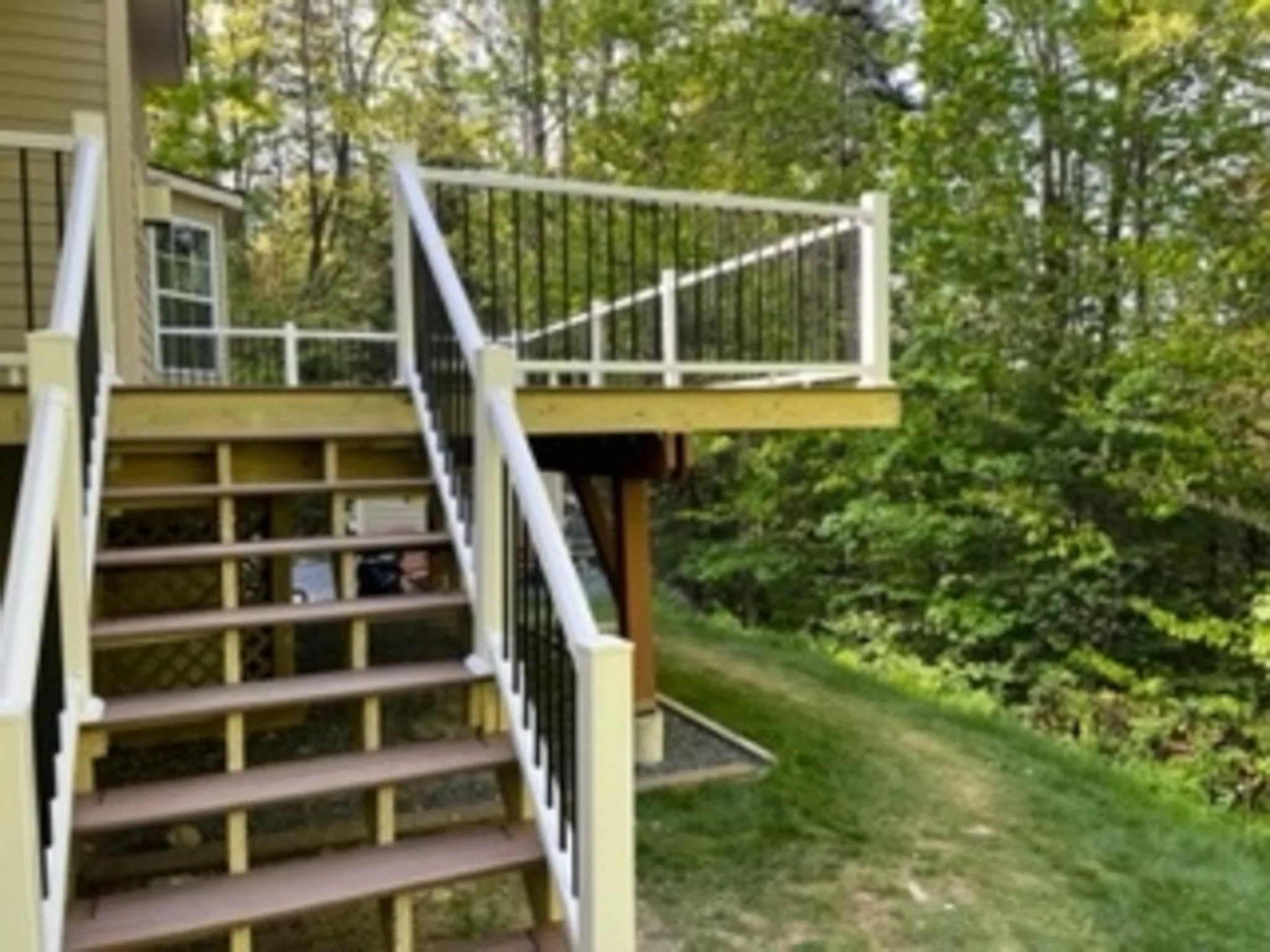 Wooden deck and staircase attached to a house with black and white railing, overlooking trees and a grassy area.