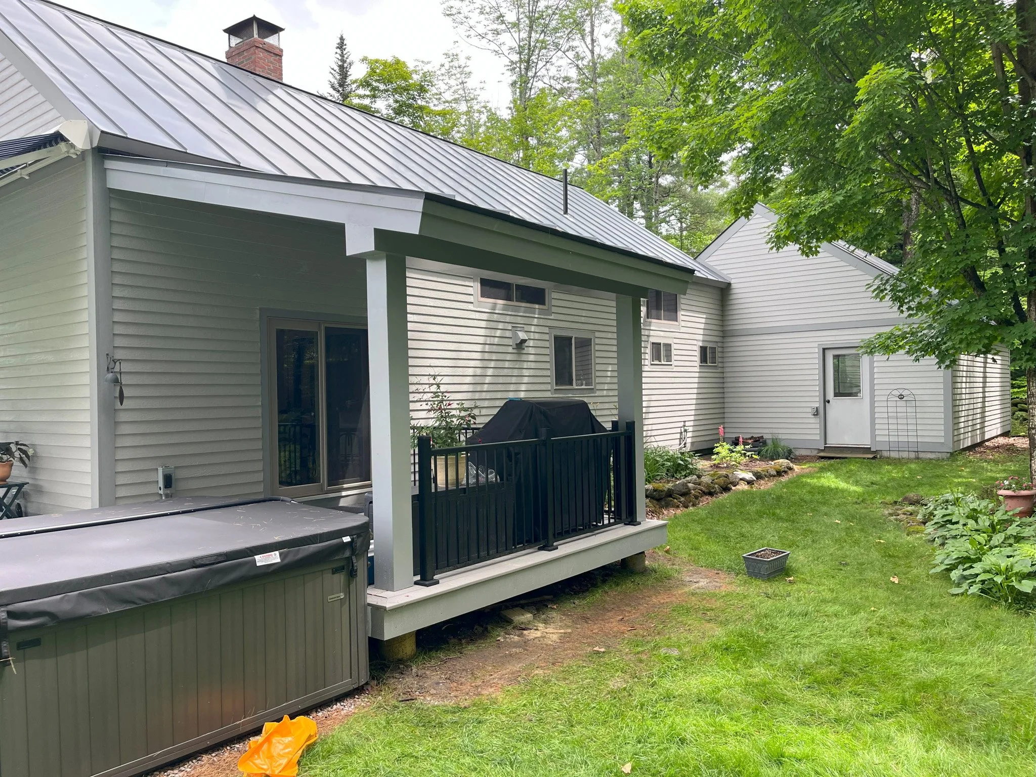 Backyard with a small deck, hot tub, and a garden with plants and rocks leading to a house with beige siding, several windows, and a white door. There is a large tree on the right side and green grass covering the yard.