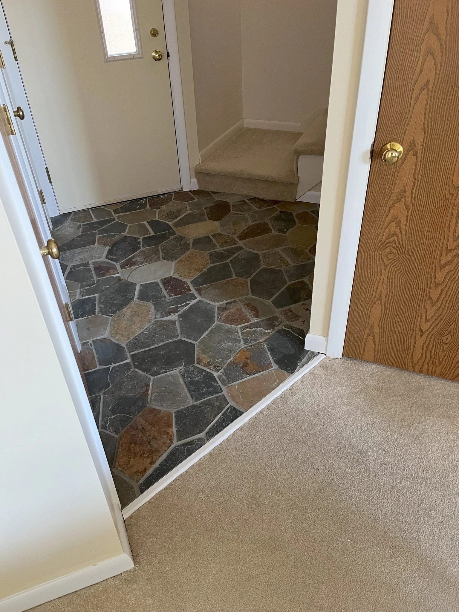 The image shows an interior view of an entryway with a transition from beige carpet to a slate tile floor near the door. There are stairs with carpeting leading upward in the corner, and a white door with a window on the left side.