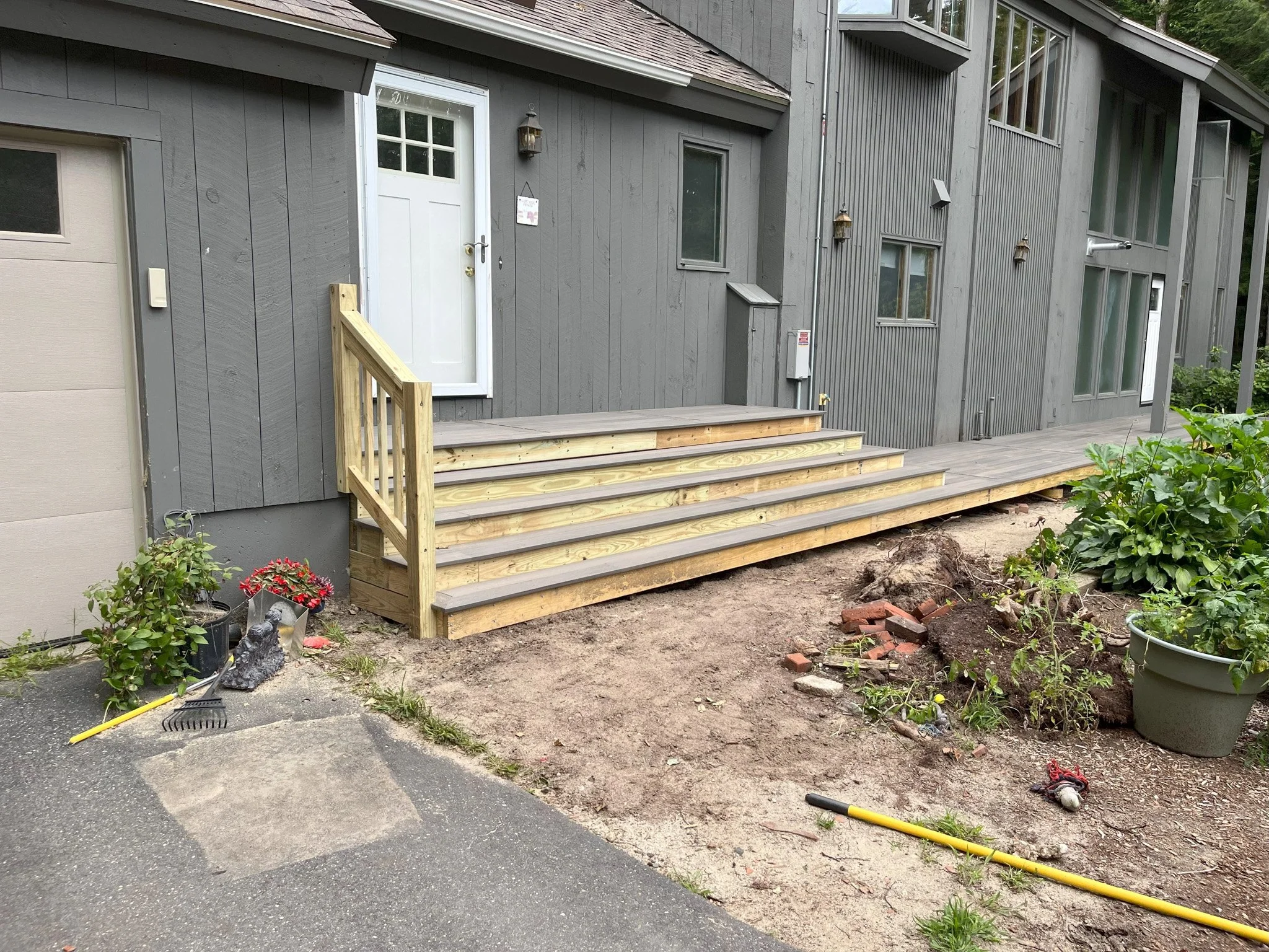 New wooden deck platform under construction in front of a gray house with a white door, surrounded by gardening tools and plants.