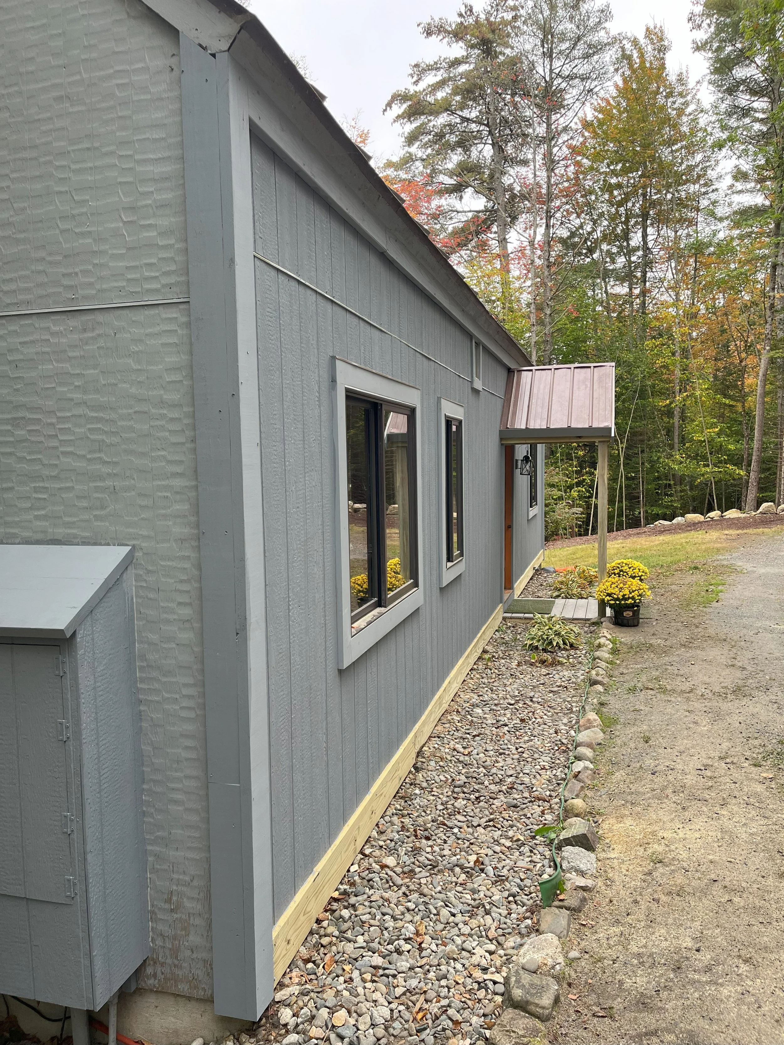 Side view of a gray house with four windows, a small porch area, and a garden with potted yellow flowers, surrounded by trees with autumn foliage.