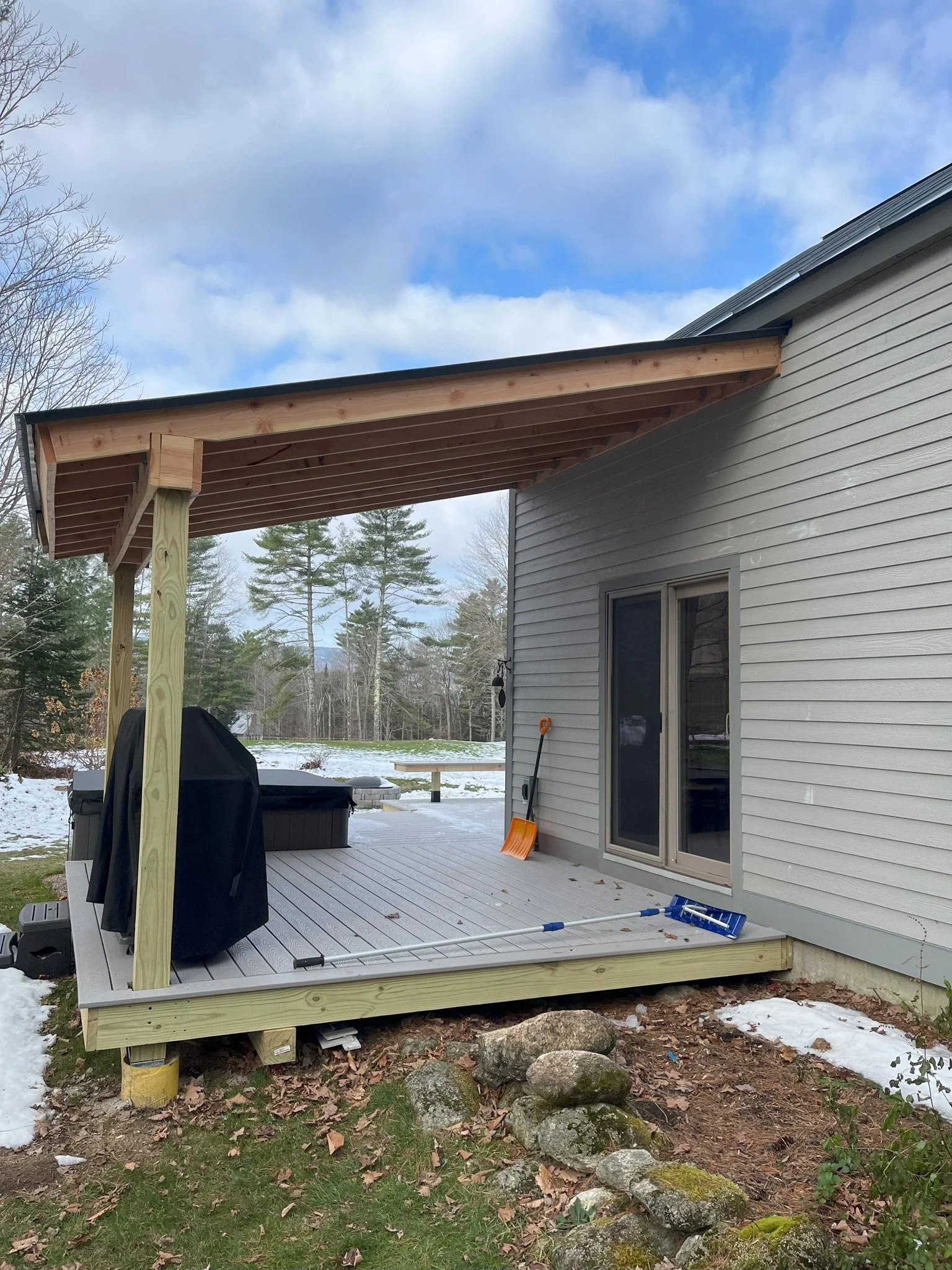 A newly built wooden deck attached to a house with sliding glass doors, surrounded by a yard with snow and trees in the background, with construction tools and supplies on the deck.