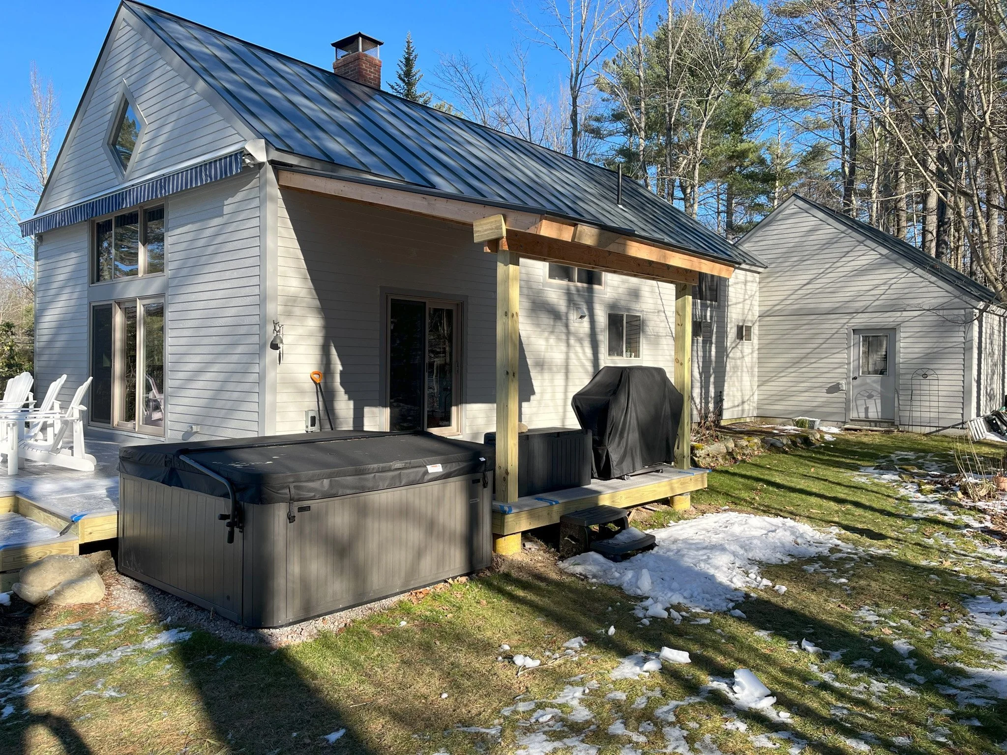 Backyard of a house with a metal roof, sliding glass door, hot tub, barbecue grill, and snow patches on the ground.