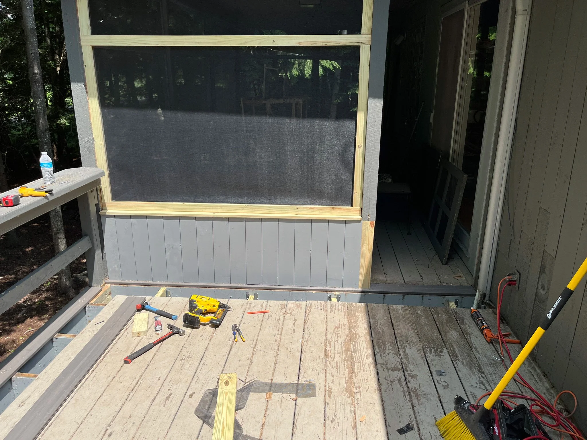 Deck construction site with tools like hammer, drill, level, and water bottle. Partially built screened porch with open doorway and construction materials.