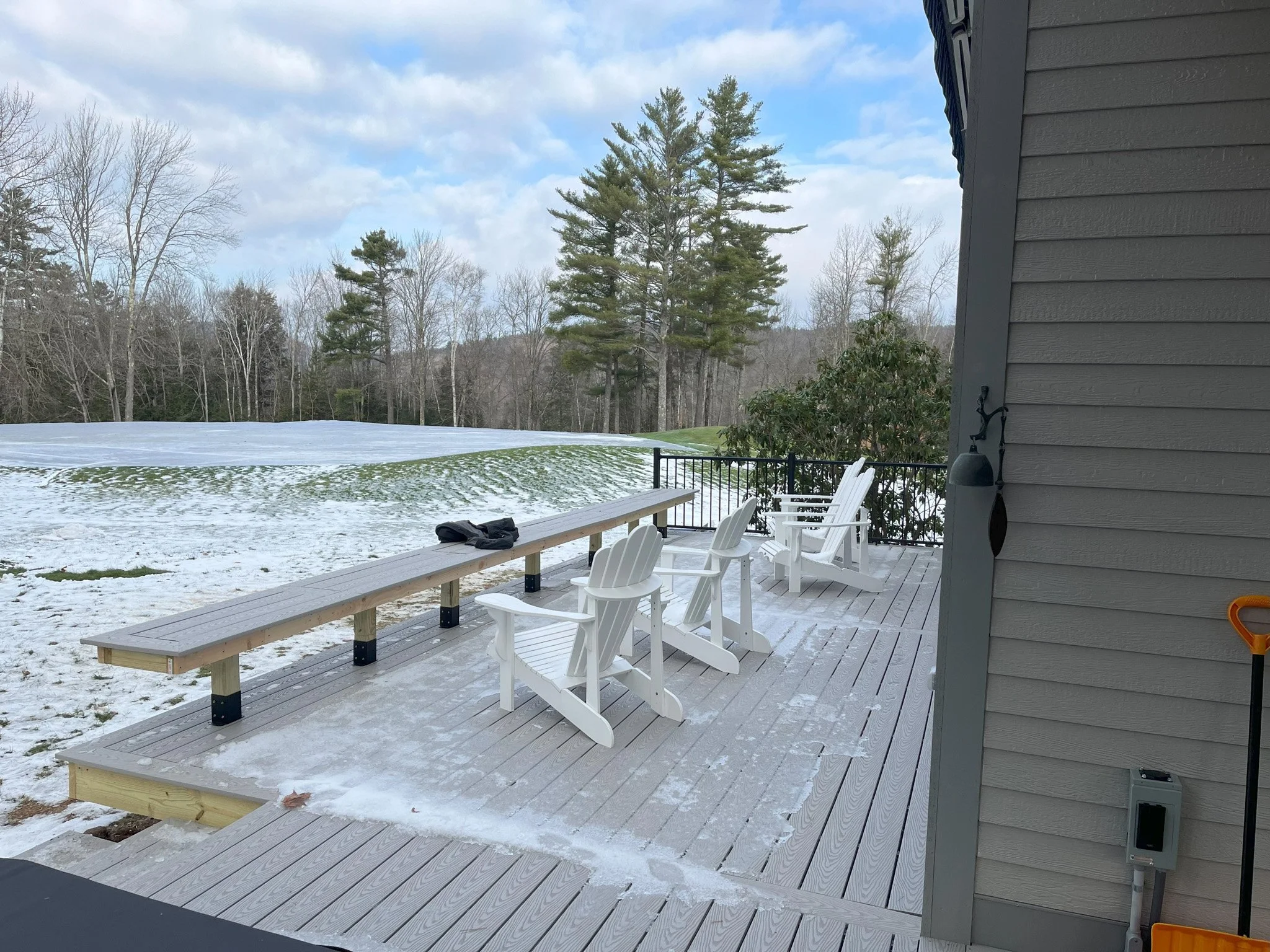 A snowy deck with white Adirondack chairs and a long wood bench overlooking a snow-covered yard and trees in the background.