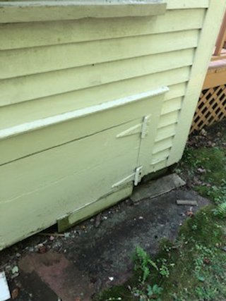 Close-up of a yellow wooden exterior wall with a damaged section near the bottom, showing chipped paint and exposed wood, next to a concrete foundation and some grass.