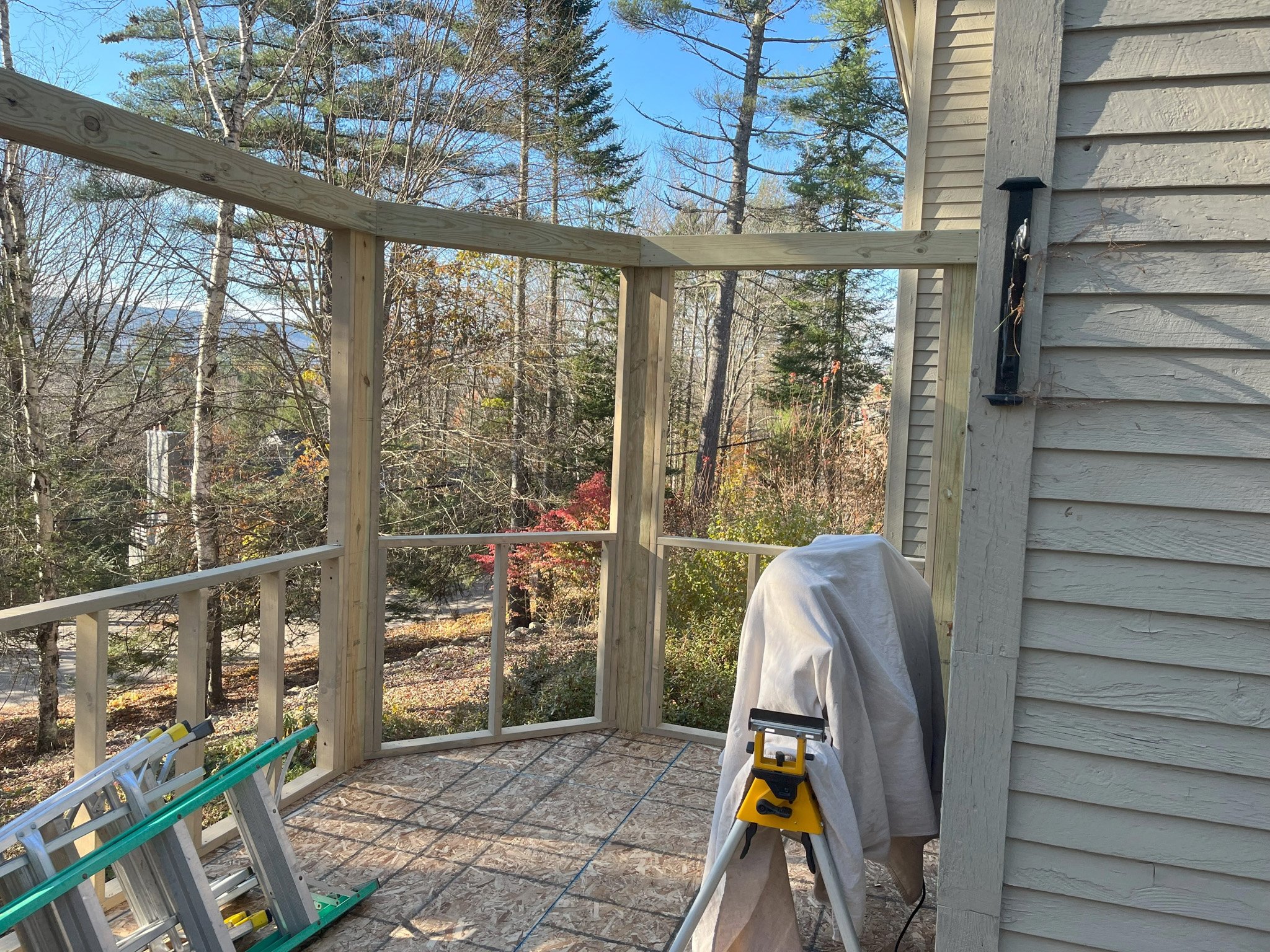 A balcony with wooden framing and a partly built railing, overlooking a wooded yard with trees and a distant view of mountains on a clear day.