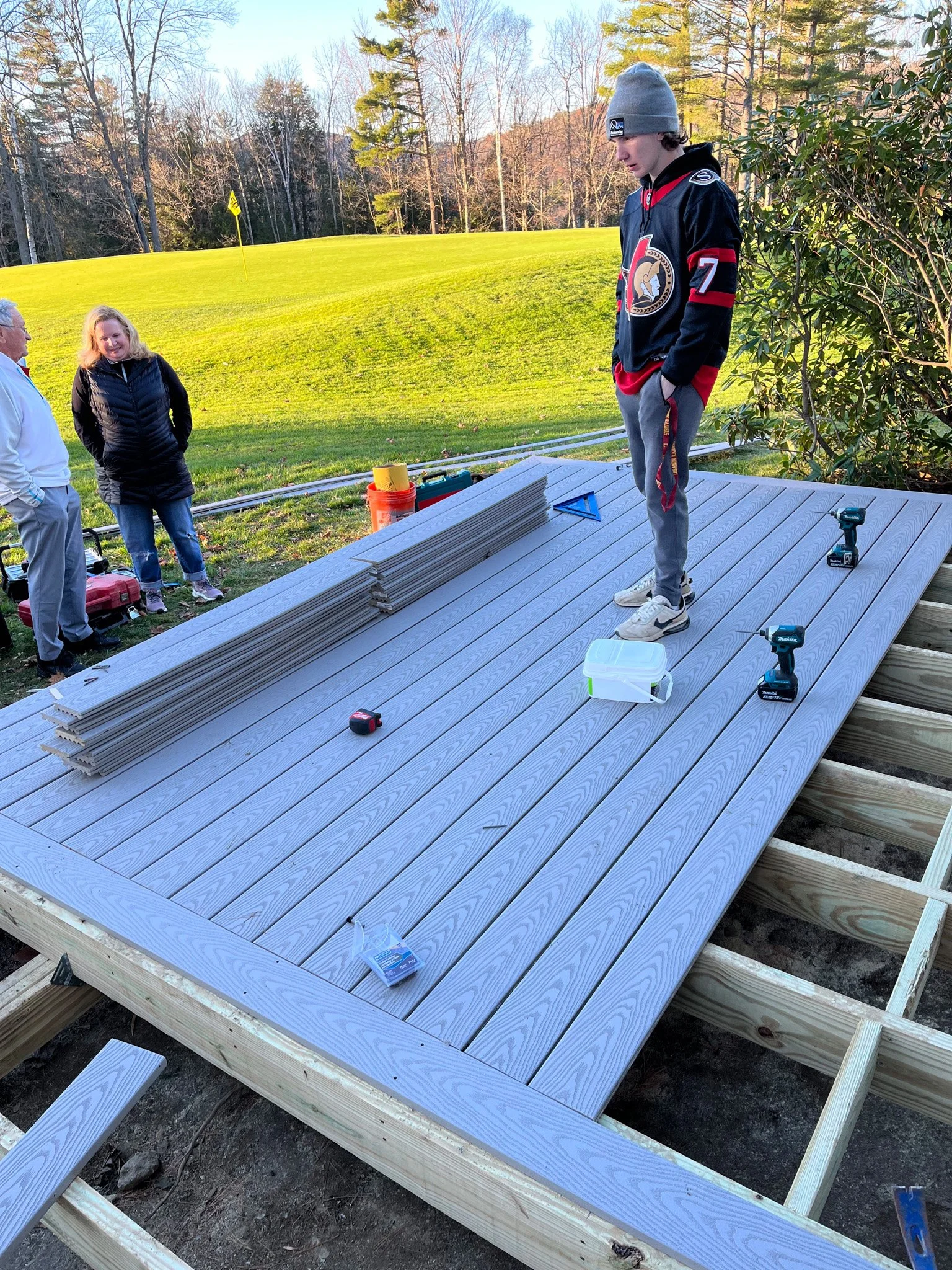 A group of people installing a wooden deck on a raised platform outdoors, with a golf course and trees in the background.