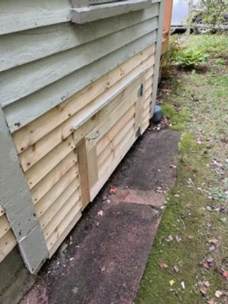 Side view of a house with new vinyl siding installed and a small door near the ground, along with a concrete walkway and grass area.