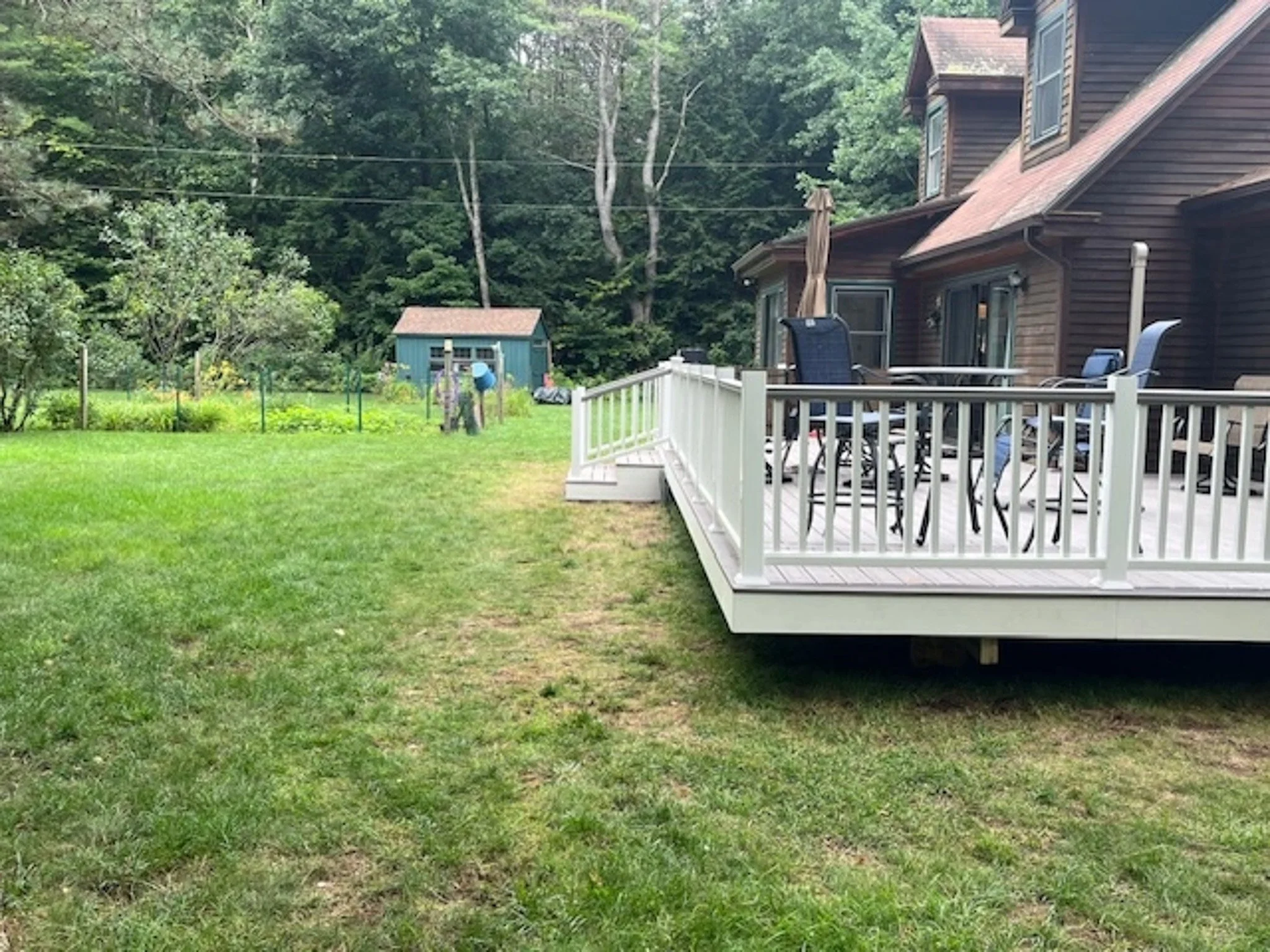 A backyard view of a house with a white deck, outdoor furniture, and a grassy lawn with trees and a shed in the background.