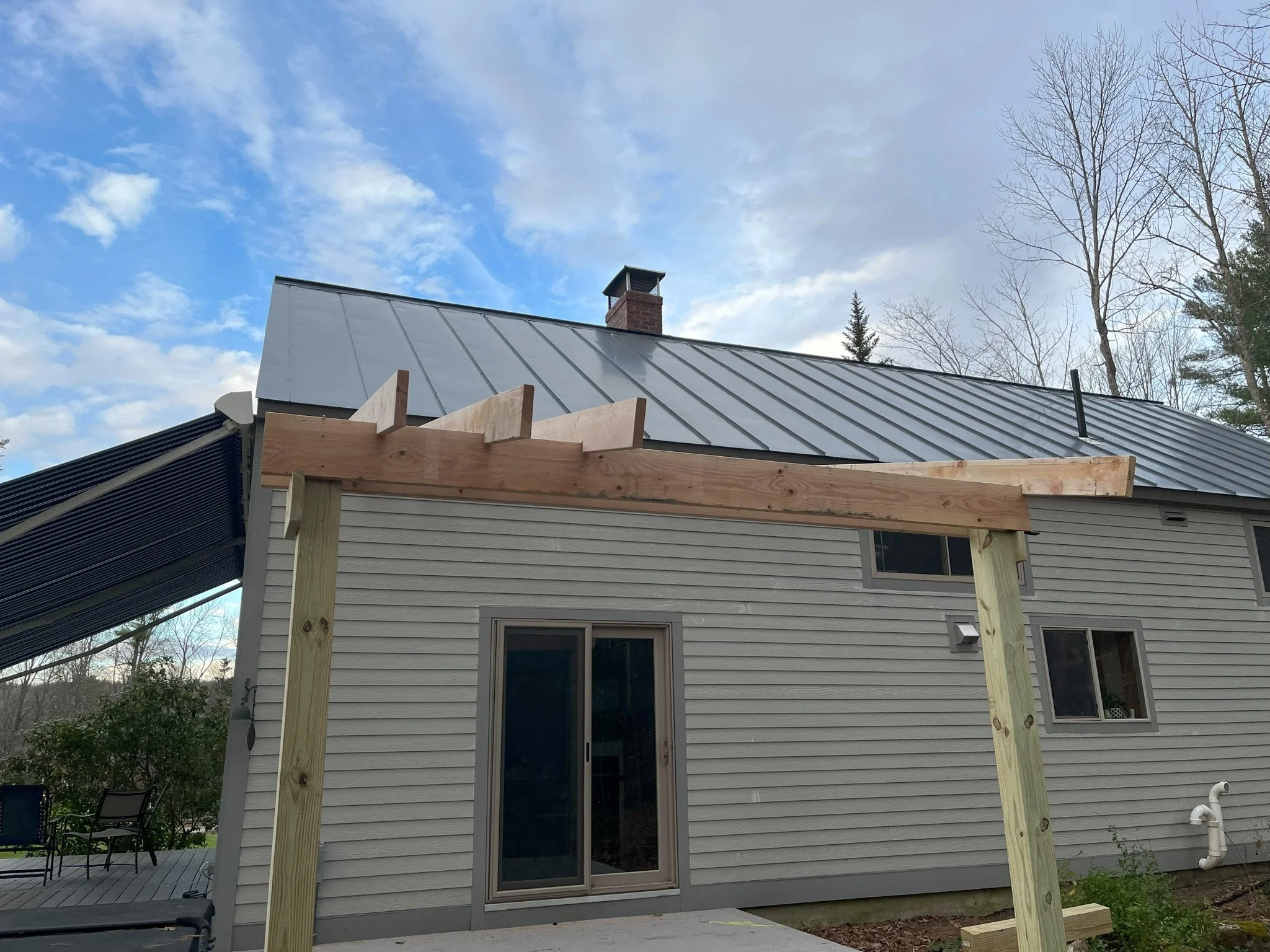 Back of a house with a new wooden pergola frame under construction, gray siding, and sliding glass door, with a sky with clouds and some trees in the background.
