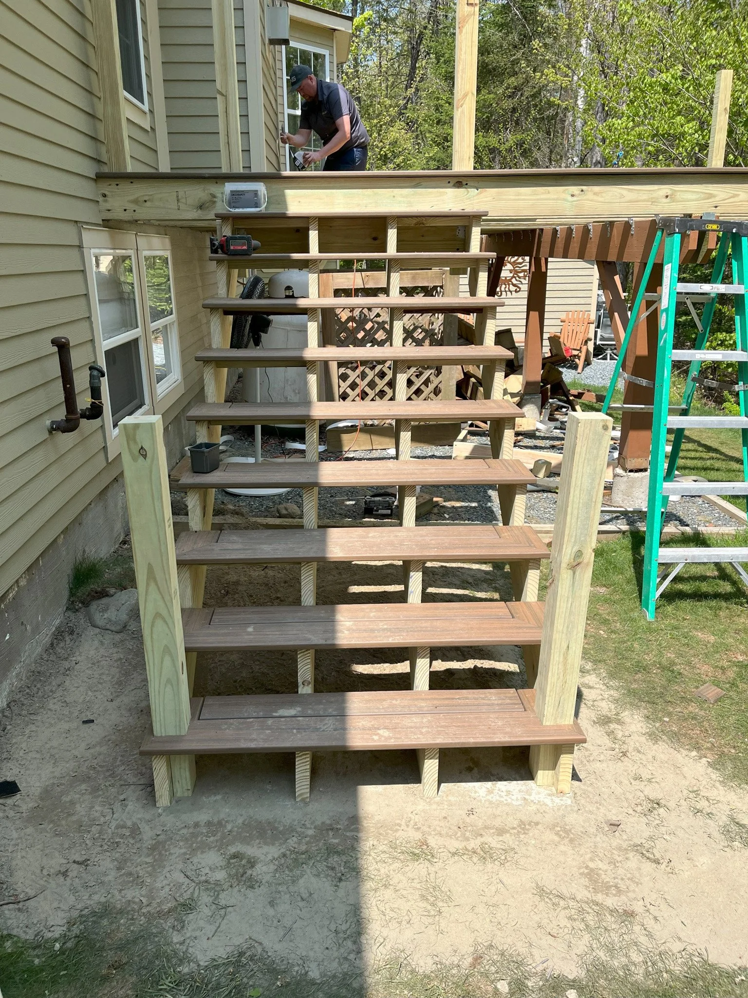 Construction of wooden stairs leading up to a deck attached to a house. A person is working on the deck, and a ladder, tools, and construction materials are visible.