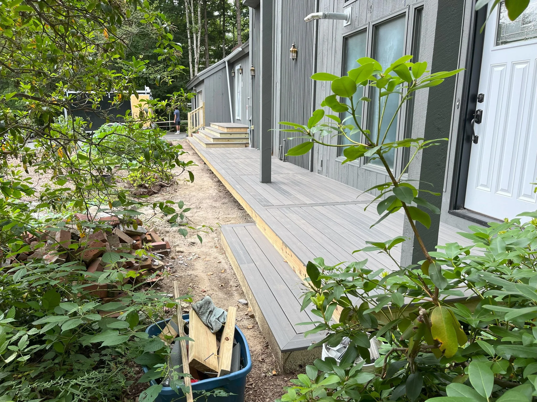 A gray wooden deck attached to a house with stairs leading up to it. There are plants and bushes in the foreground, and a person walking in the background.