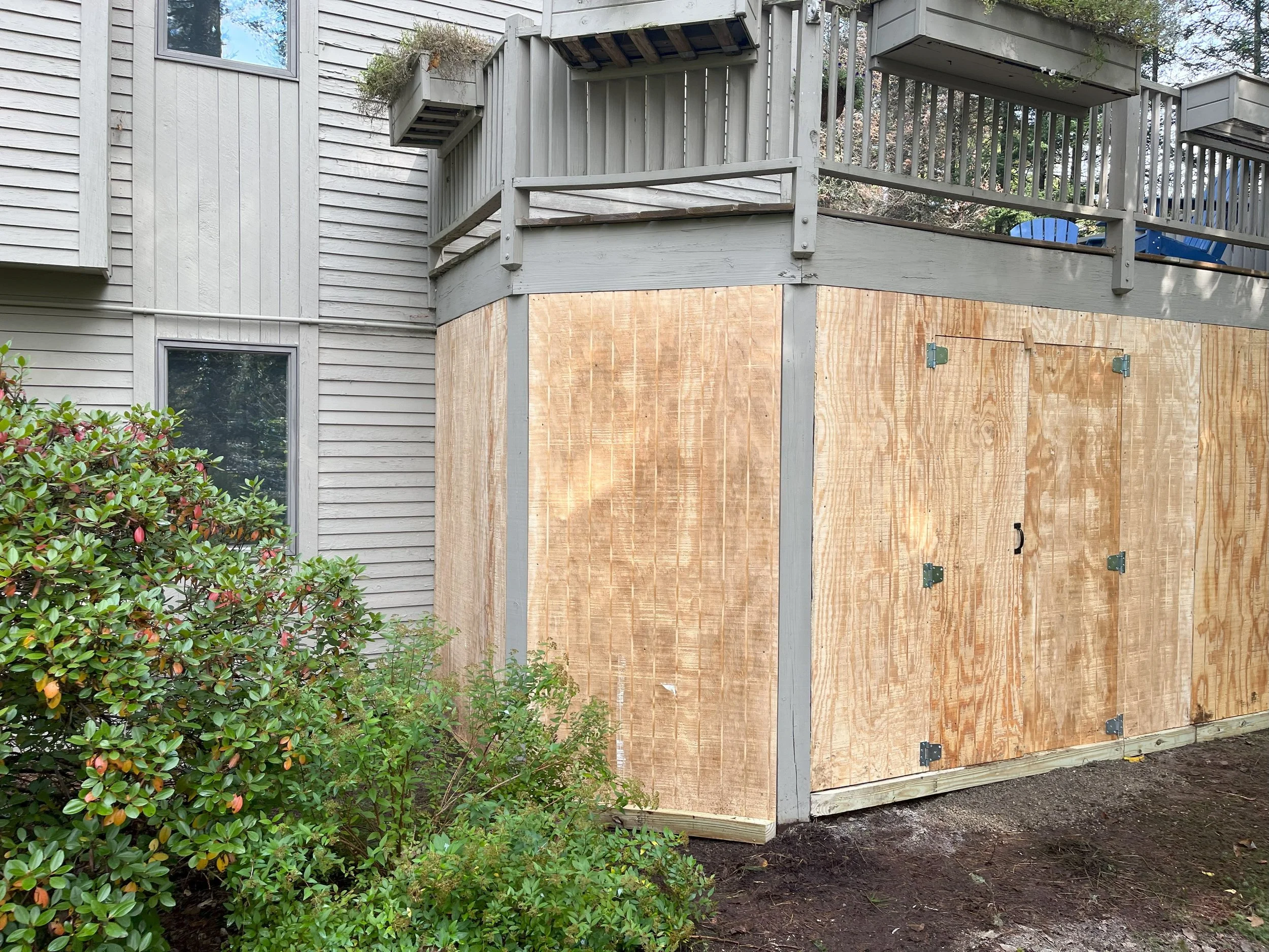 Side view of a house with a new wooden deck enclosure under construction, featuring plywood walls and a railing on top. There are plants and bushes in the foreground.