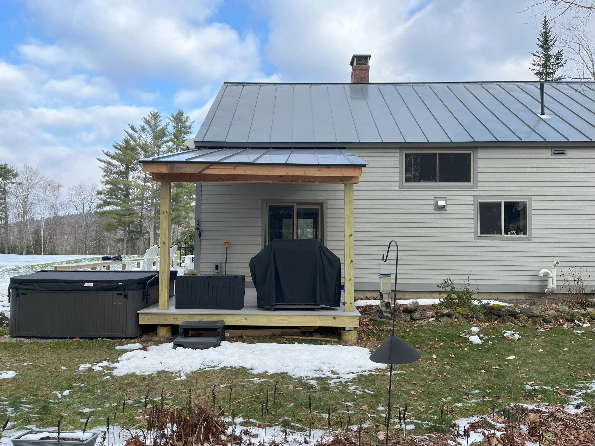 Backyard of a house with a small deck, hot tub, grill, snow patches, and trees in the background.