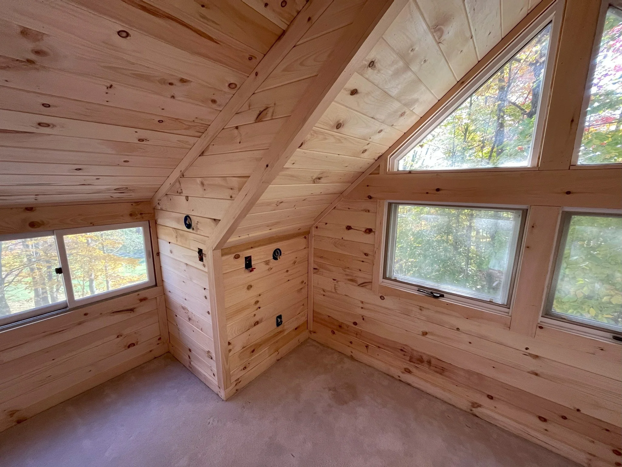 Interior of an unfinished wooden room with multiple windows, showing wood paneling on the walls and ceiling, and an unfinished wall with electrical outlets and wiring.