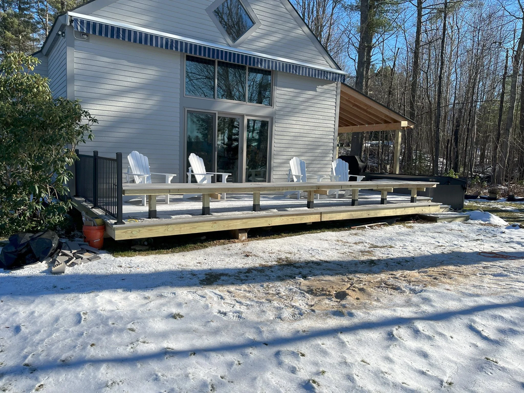 A house with a wooden deck, four white Adirondack chairs, and a snow-covered yard during winter. There are trees in the background and a partially covered outdoor area with a grill.