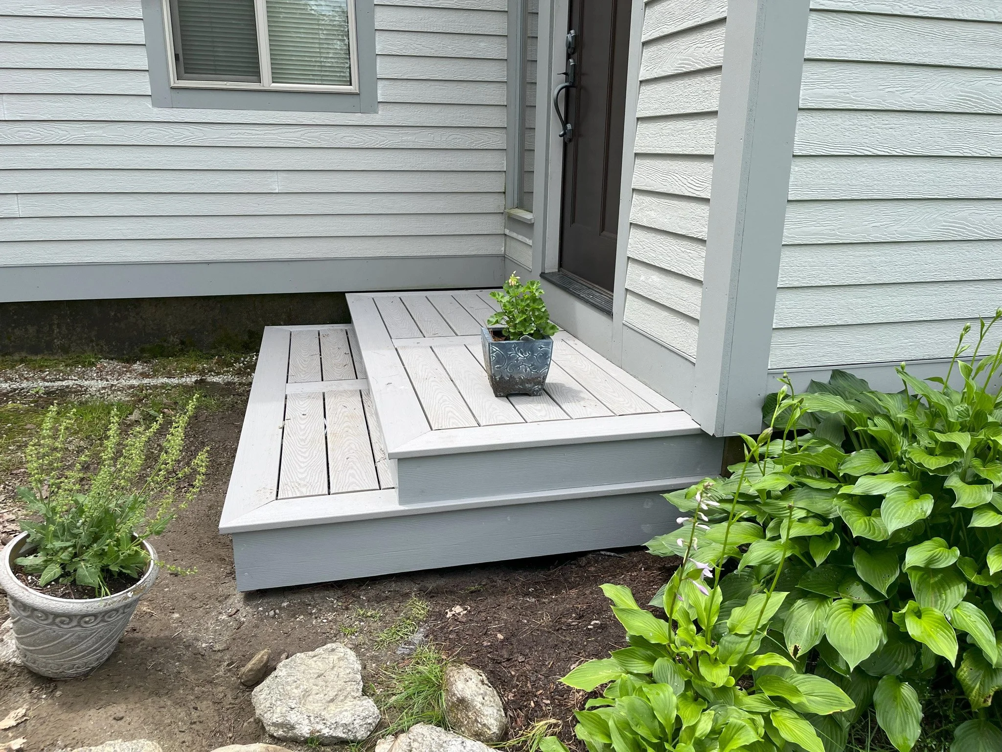 Small wooden porch with gray painted steps and a black front door with a decorative strap handle, surrounded by green plants and potted flowers.