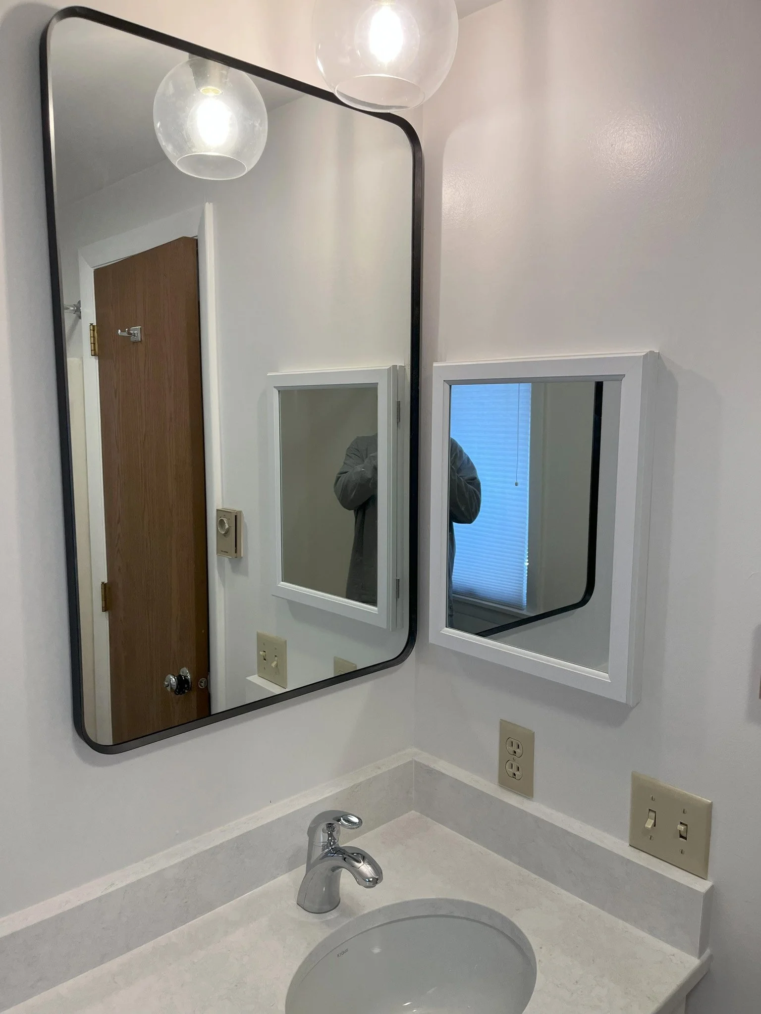 Bathroom with two mirrors, one large with rounded corners and a smaller rectangular one, white walls, a countertop with sink, and visible electrical outlets. A reflection shows a person taking the photo, and a window with closed blinds is also visibl