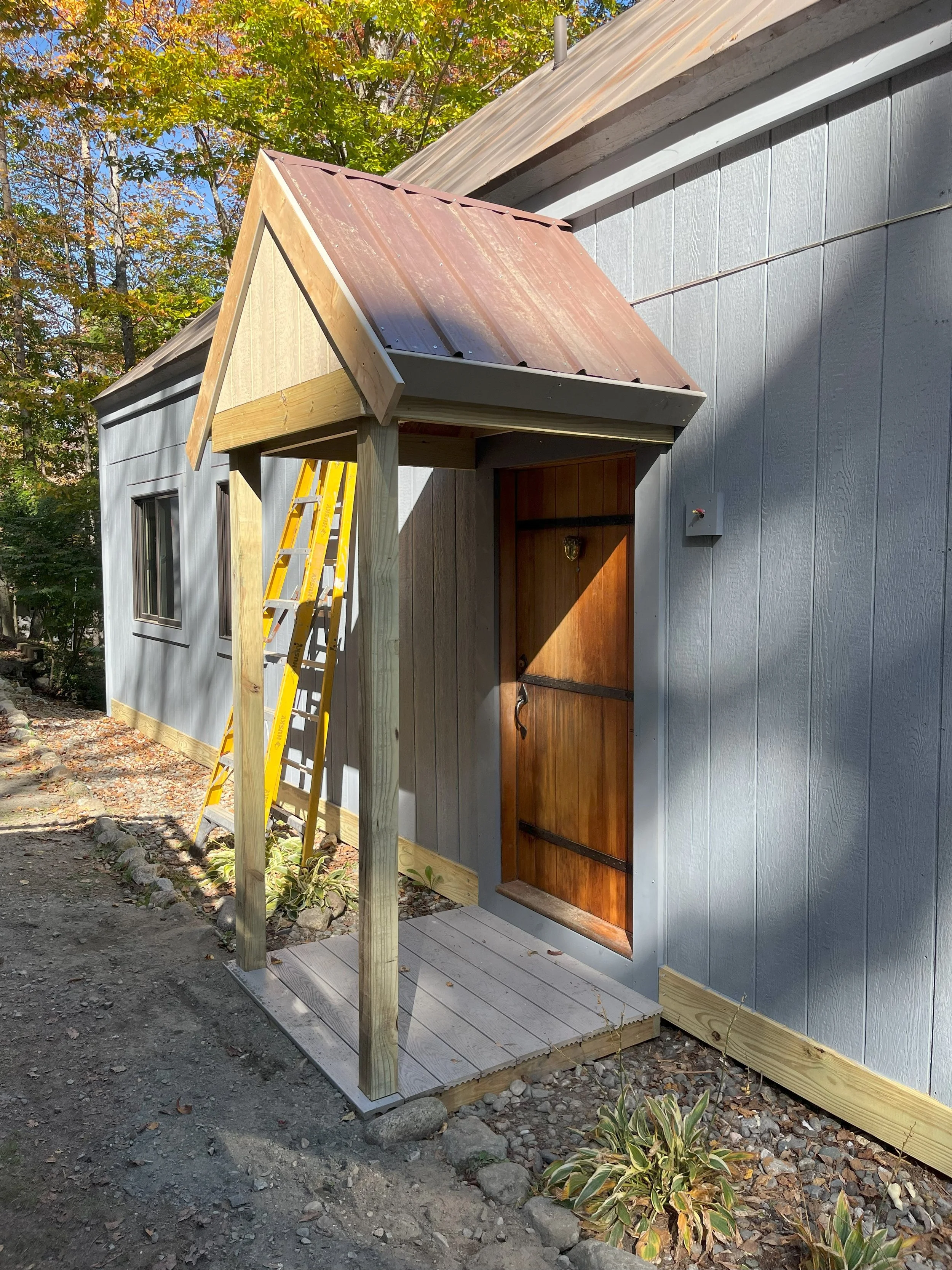 Small wooden structure with a pitched metal roof, a wooden door, and a small platform in front, attached to a gray building with siding.