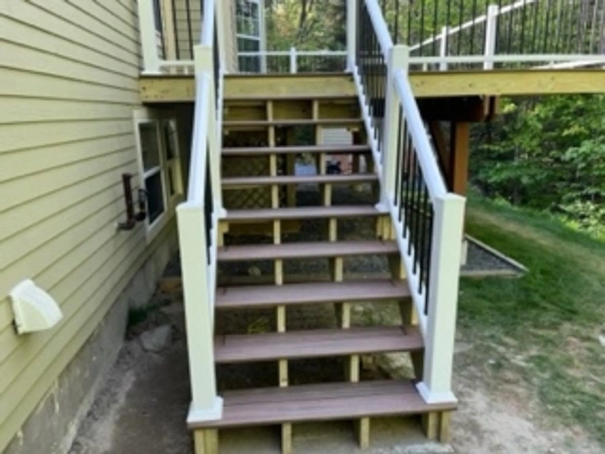 New wooden outdoor staircase with white railings attached to a yellow house, leading to a deck. Surrounding greenery visible.