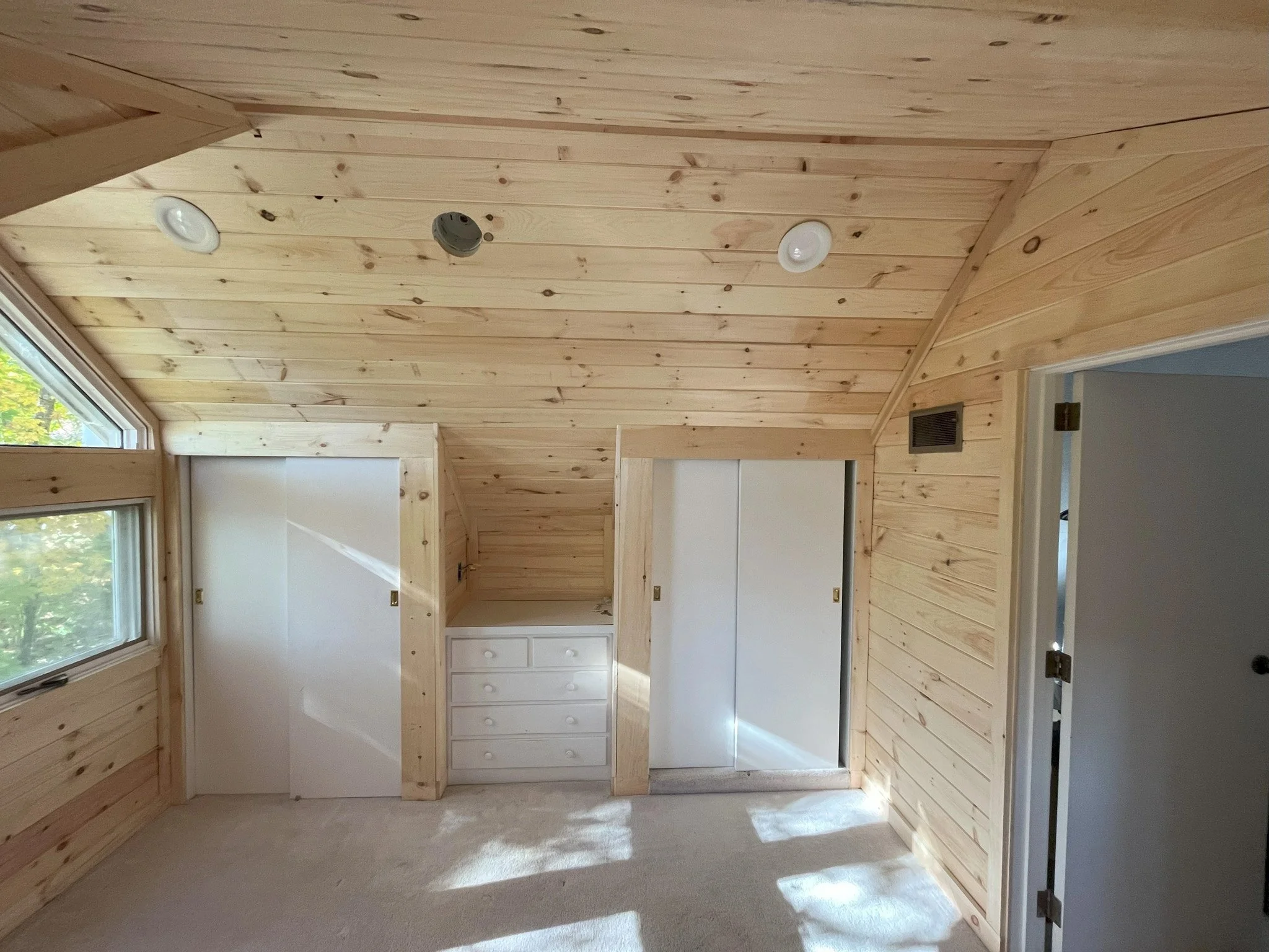 Interior of a room with wood-paneled walls and ceiling, featuring two closet doors and a small set of white drawers, with a window on the left letting in natural light.