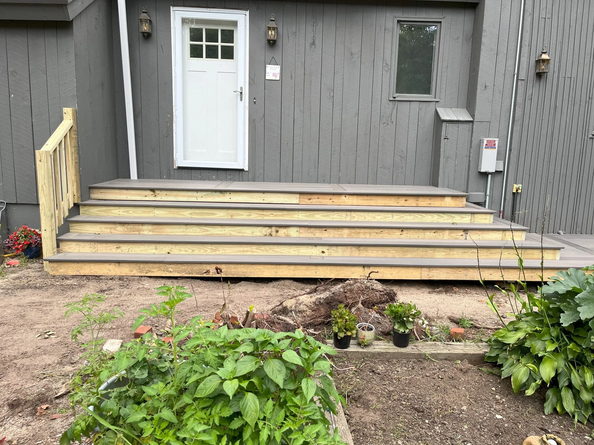 Wooden deck under construction in front of a gray house with a white door, with potted plants and garden greenery in the foreground.