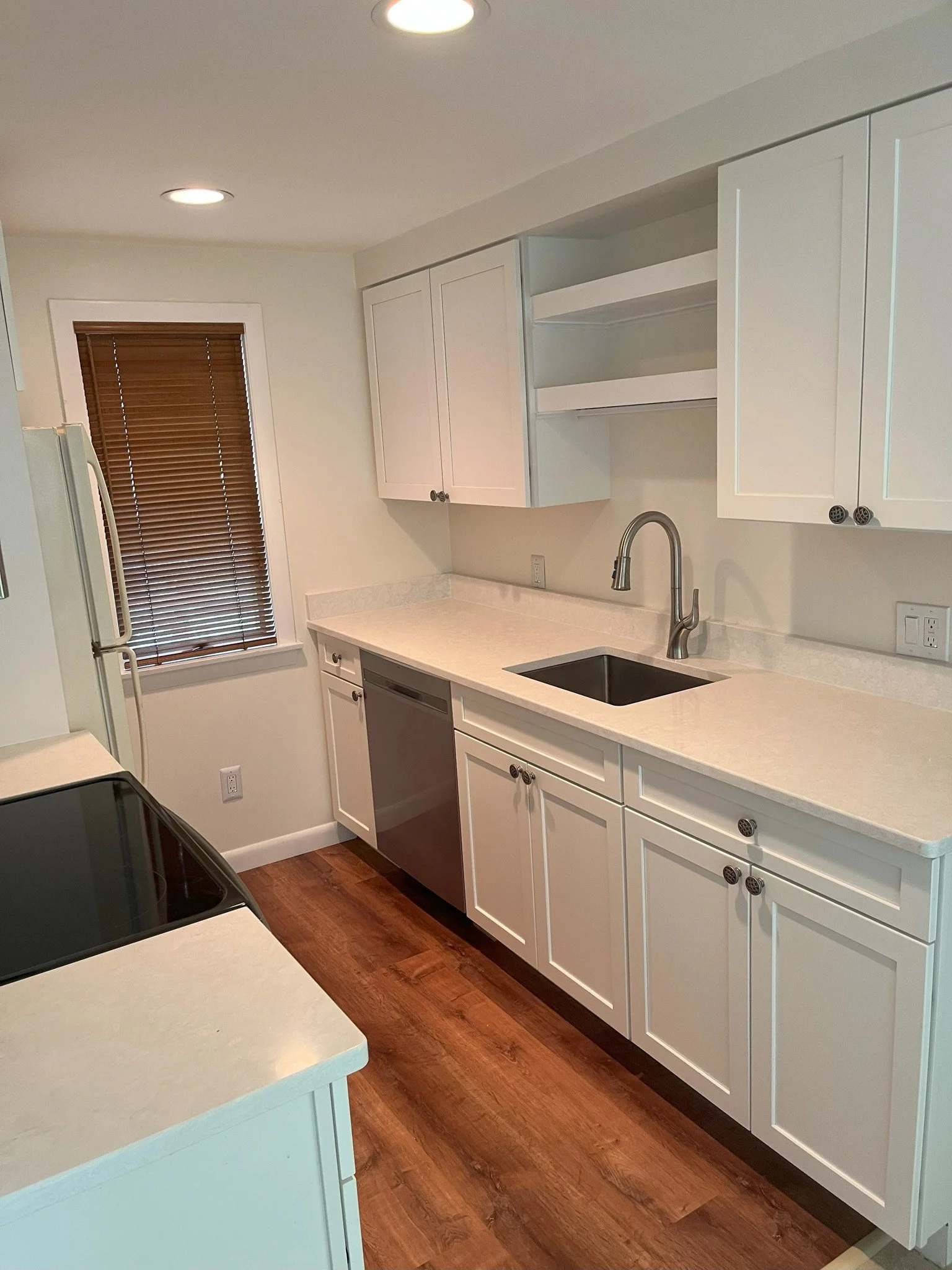 A modern kitchen with white cabinets, a white countertop, a black sink, a dishwashing machine, a stove, a window with brown blinds, and wooden flooring.