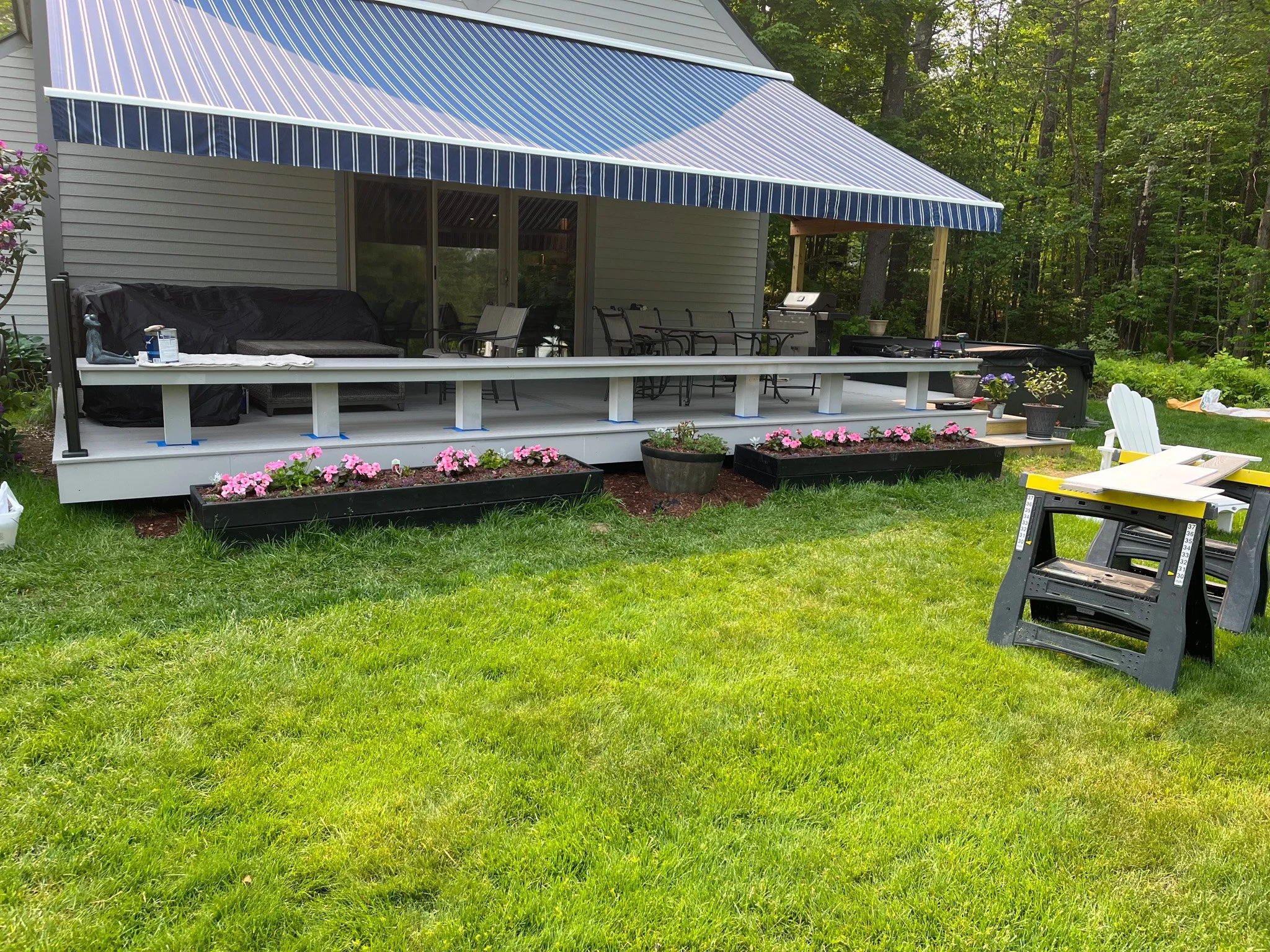 A backyard patio with a decorated wooden deck, a blue and white striped awning, flower boxes with pink flowers, a black grill, and outdoor furniture situated in a grassy area with trees in the background.