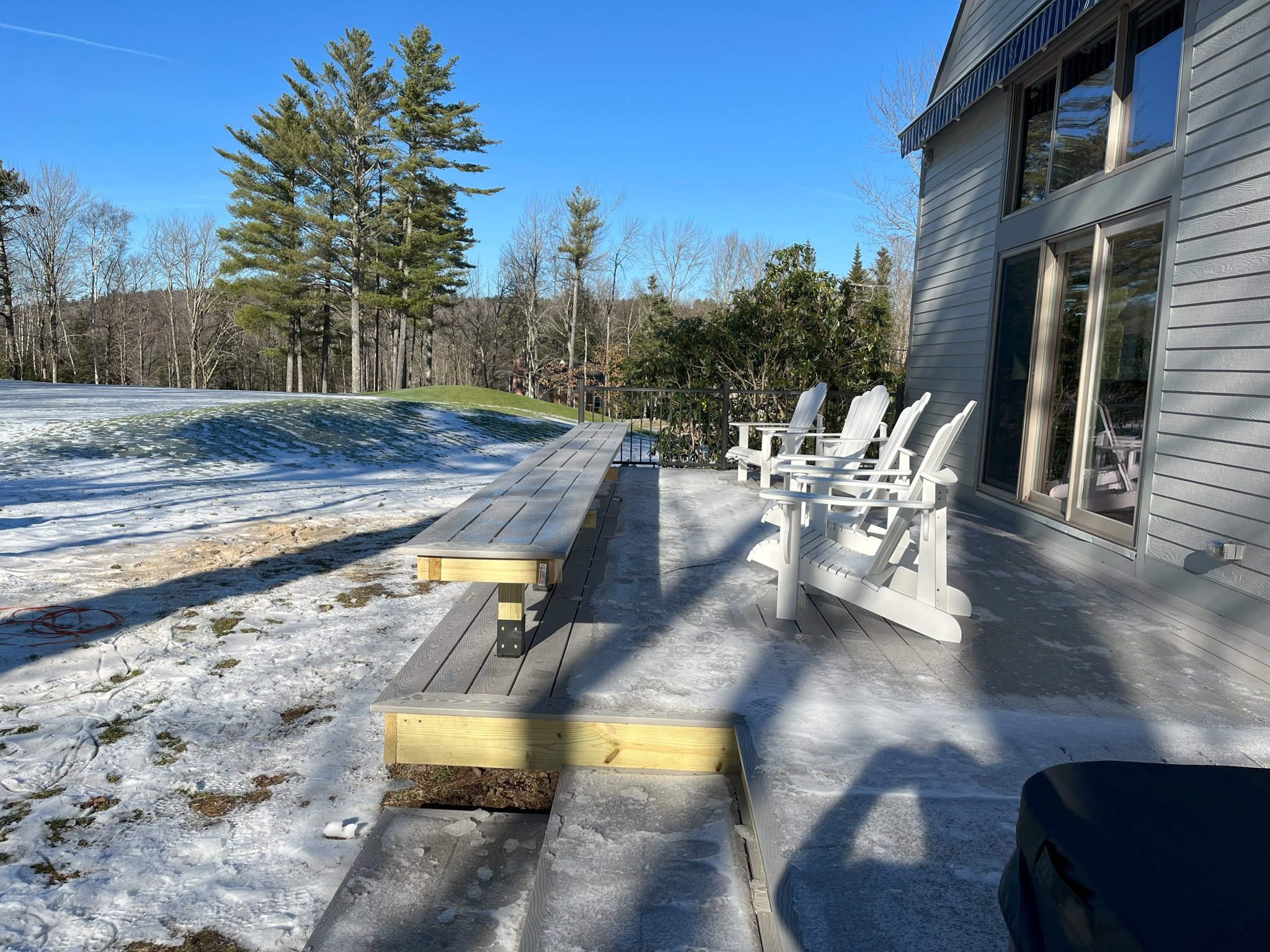 Backyard deck with four white Adirondack chairs on icy surface, partial snow-covered lawn, large pine trees, house with gray siding and sliding doors, blue sky, and bare trees in the background.