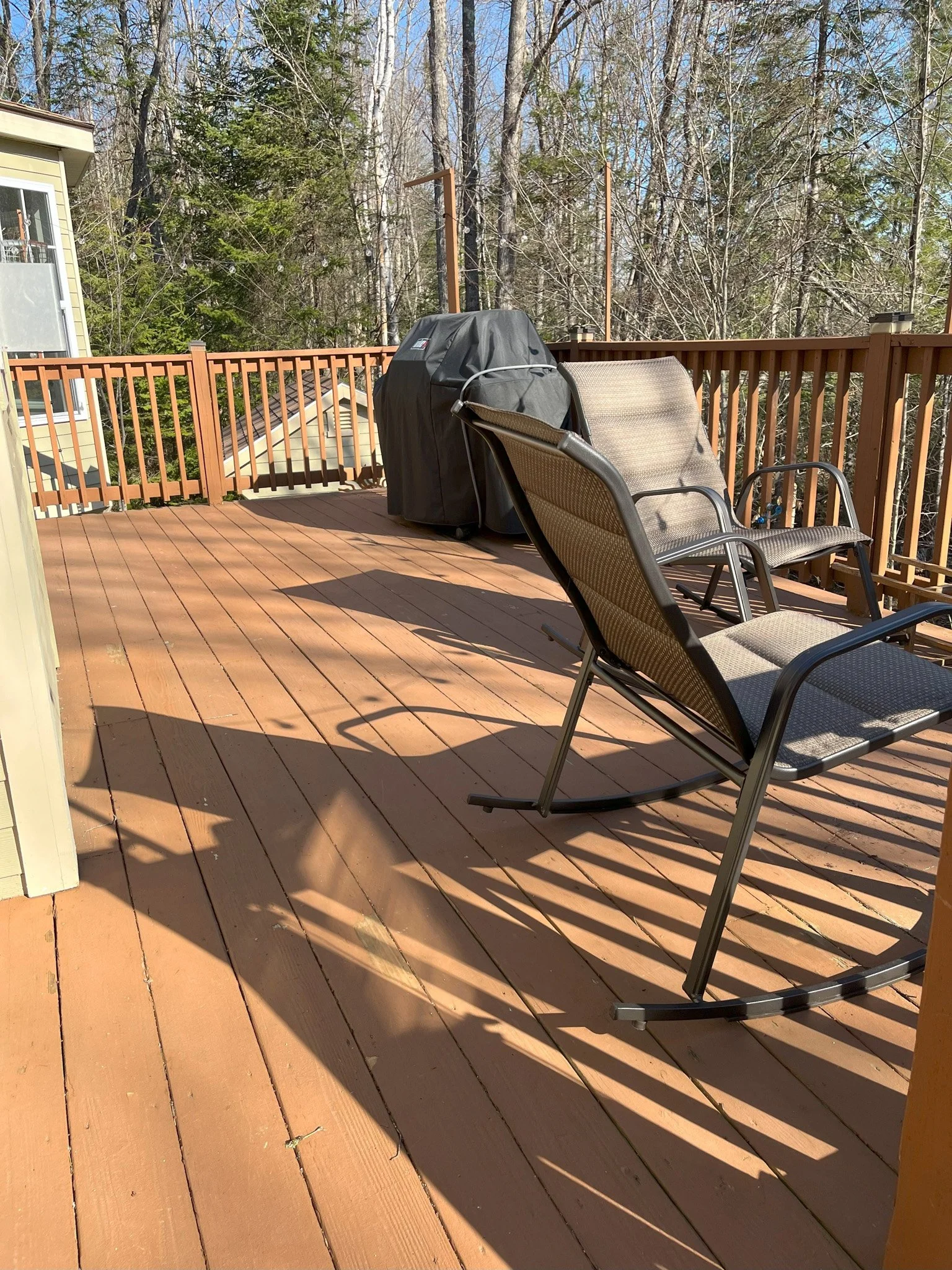 Sunlit wooden deck with patio furniture, including two rocking chairs and a recliner, surrounded by fencing and adjacent to a house, with trees and clear sky in the background.