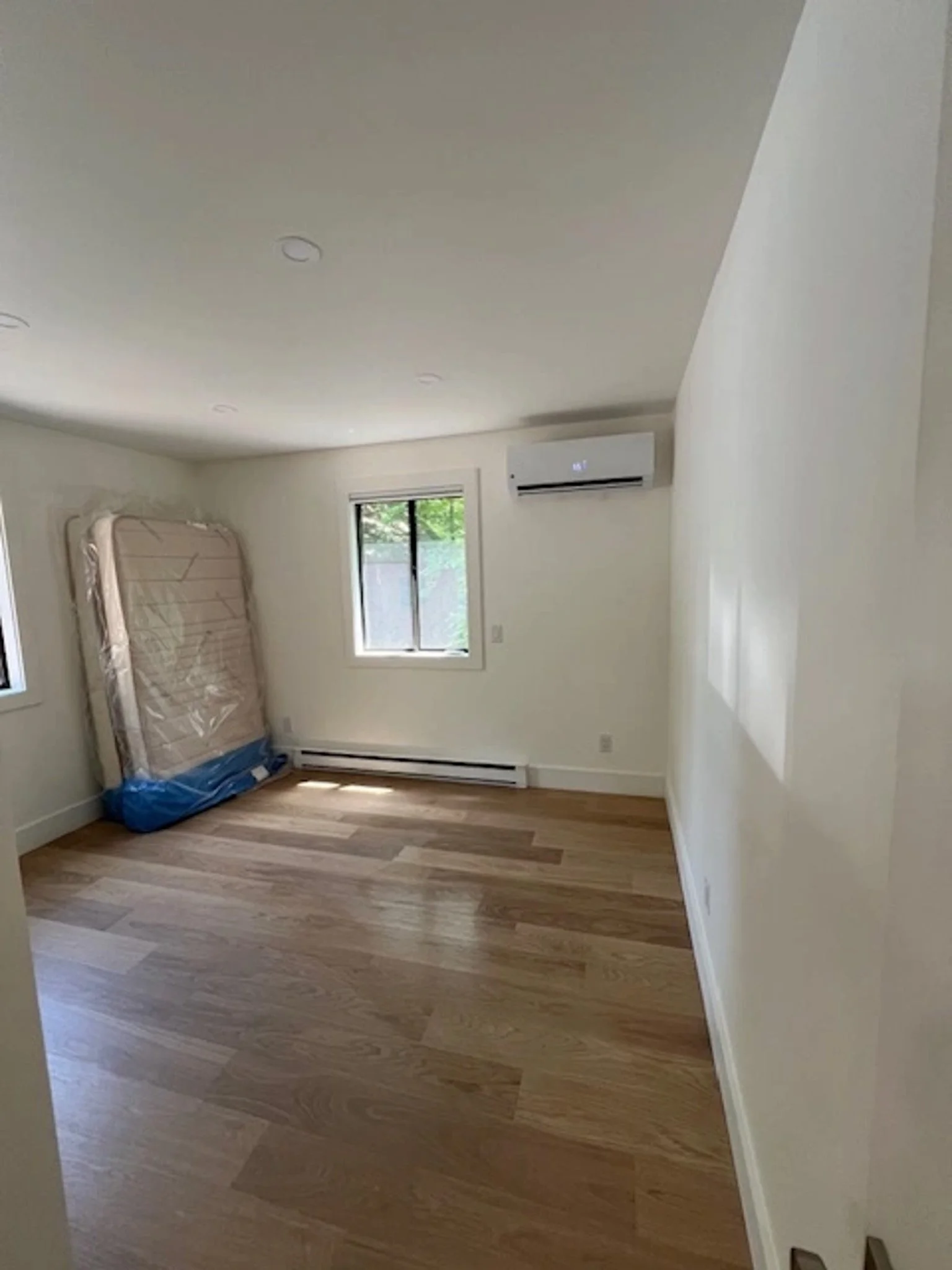 Empty room with hardwood floors, white walls, two windows, an air conditioning unit, and a partially visible mattress leaning against the wall.