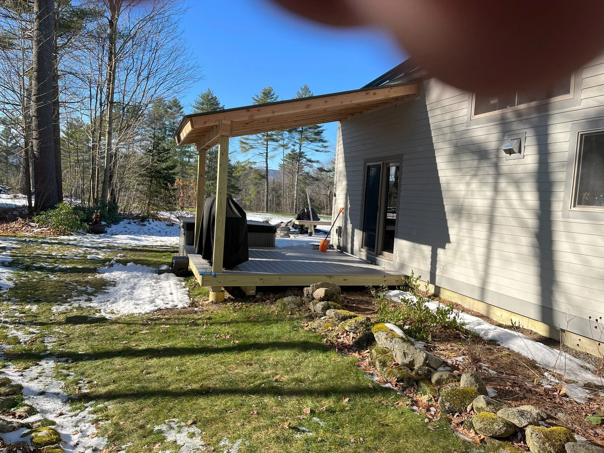 Backyard patio with partial cover, deck, grill, snow patches, and trees in the background.