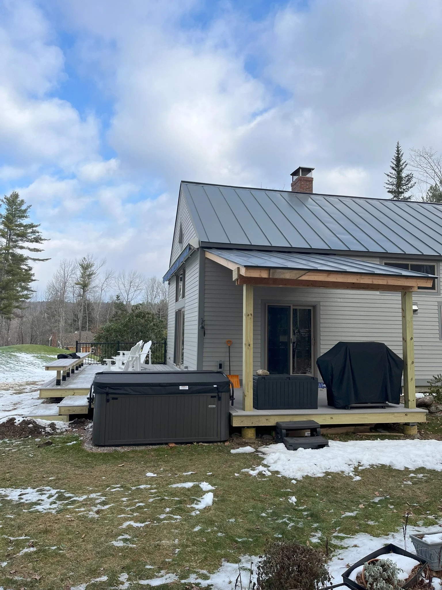 Backyard of a house with a metal roof, small deck, outdoor furniture, a grill, and hot tub, with snow on the grass and trees in the background.