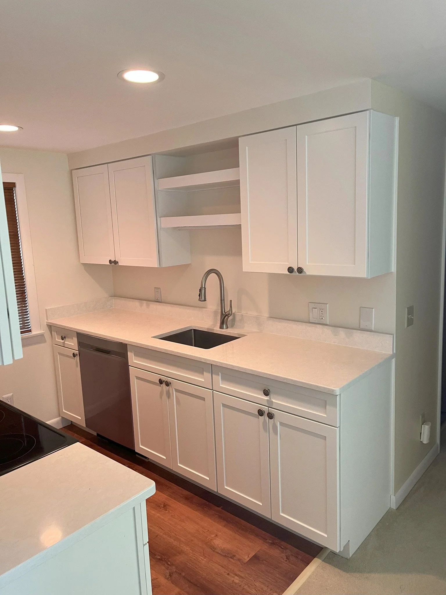 Modern kitchen with white cabinets, a sink with a high-arc faucet, a built-in dishwasher, and open shelving above the countertop. Recessed lighting in the ceiling.