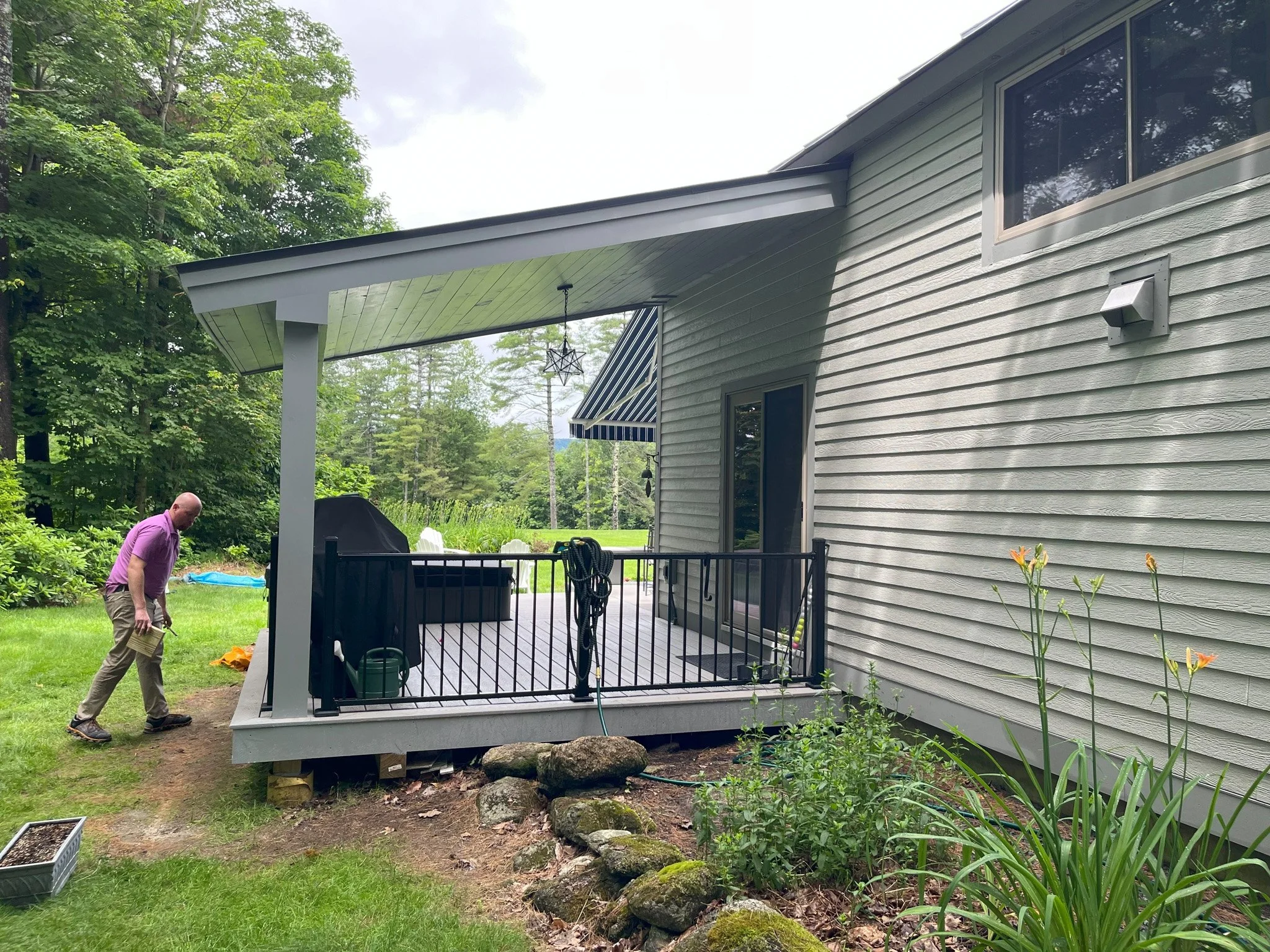 A man in a purple shirt and beige pants walking near a newly constructed backyard deck attached to a house with beige siding. The deck has black railings and outdoor furniture, with a garden and trees in the background.