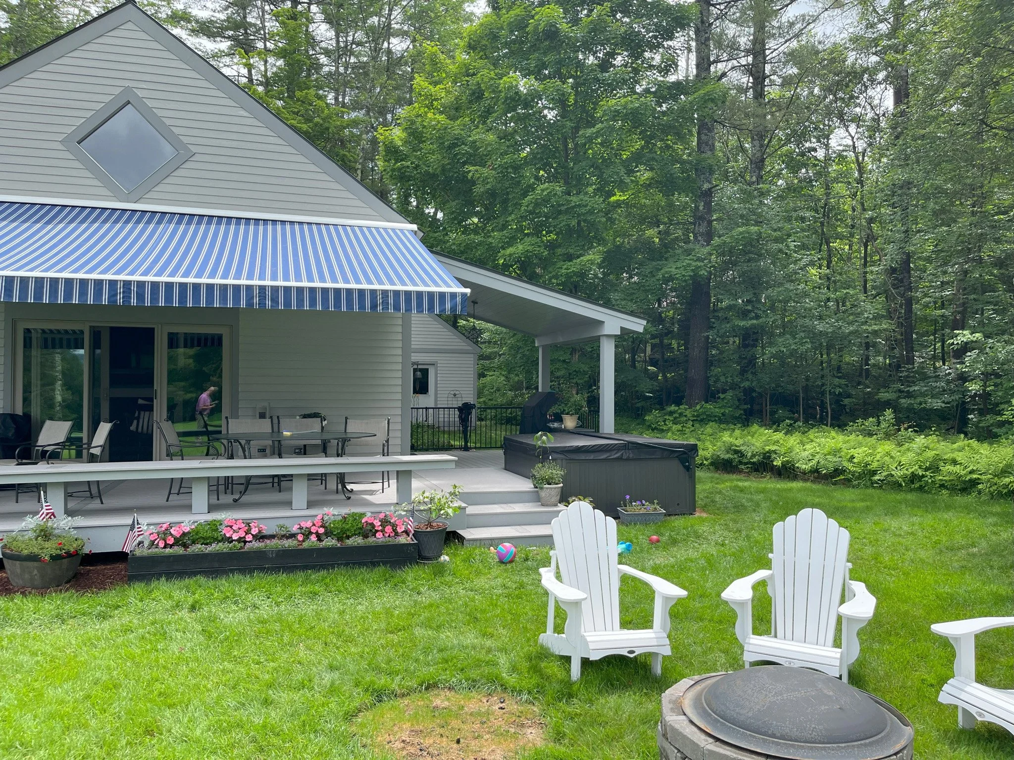 Backyard patio with outdoor chairs, a table, potted plants, a covered hot tub, and a sliding glass door with an awning. There are trees and greenery in the background.