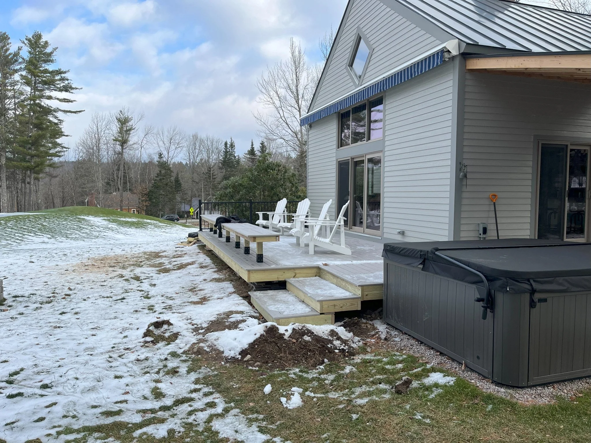 Backyard patio with a wooden deck with steps, white Adirondack chairs, a hot tub, and a snow-covered lawn surrounding the deck.