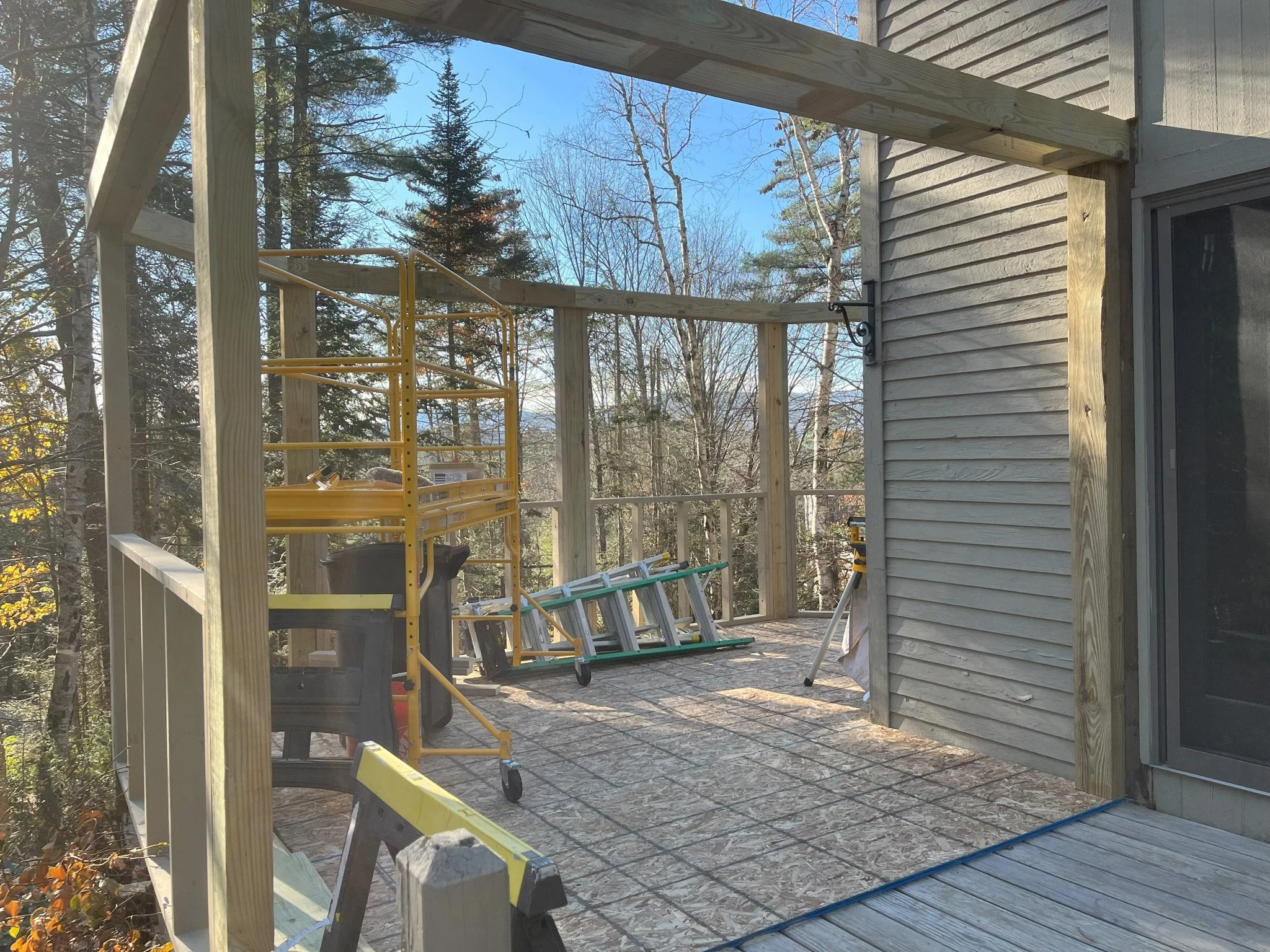 Construction of a new porch on the side of a house, with wooden framing and tools including a ladder and scaffolding, amid trees with fall foliage in the background.