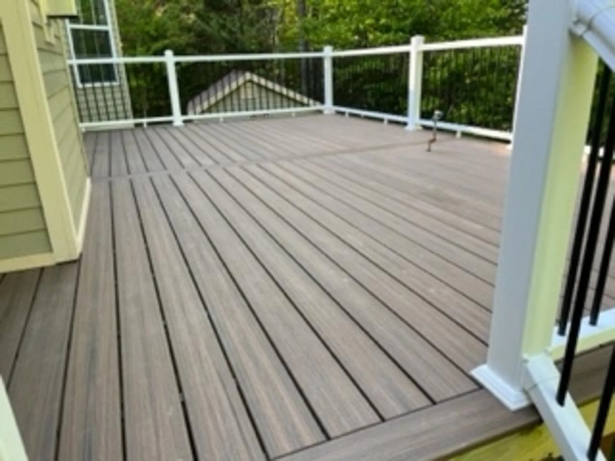 Empty wooden deck with white railing, attached to a yellow house, surrounded by green trees in the background.