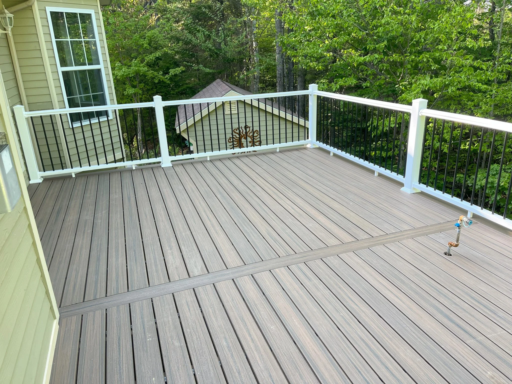 Empty outdoor wooden deck with white and black railing, connected to a yellow house, surrounded by green trees.